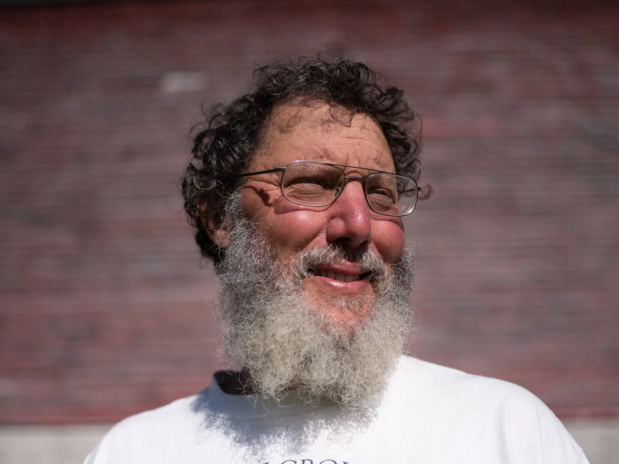 Close-up of a smiling Vermont farmer with curly hair, glasses, and a beard, wearing a white top, against a blurred background.
