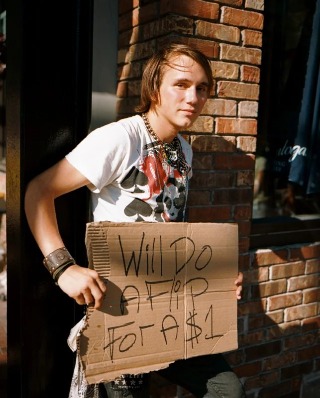 Young man with shoulder-length hair wearing a graphic T-shirt, standing against a brick wall, holding a cardboard sign that reads "Will do A for A$1."