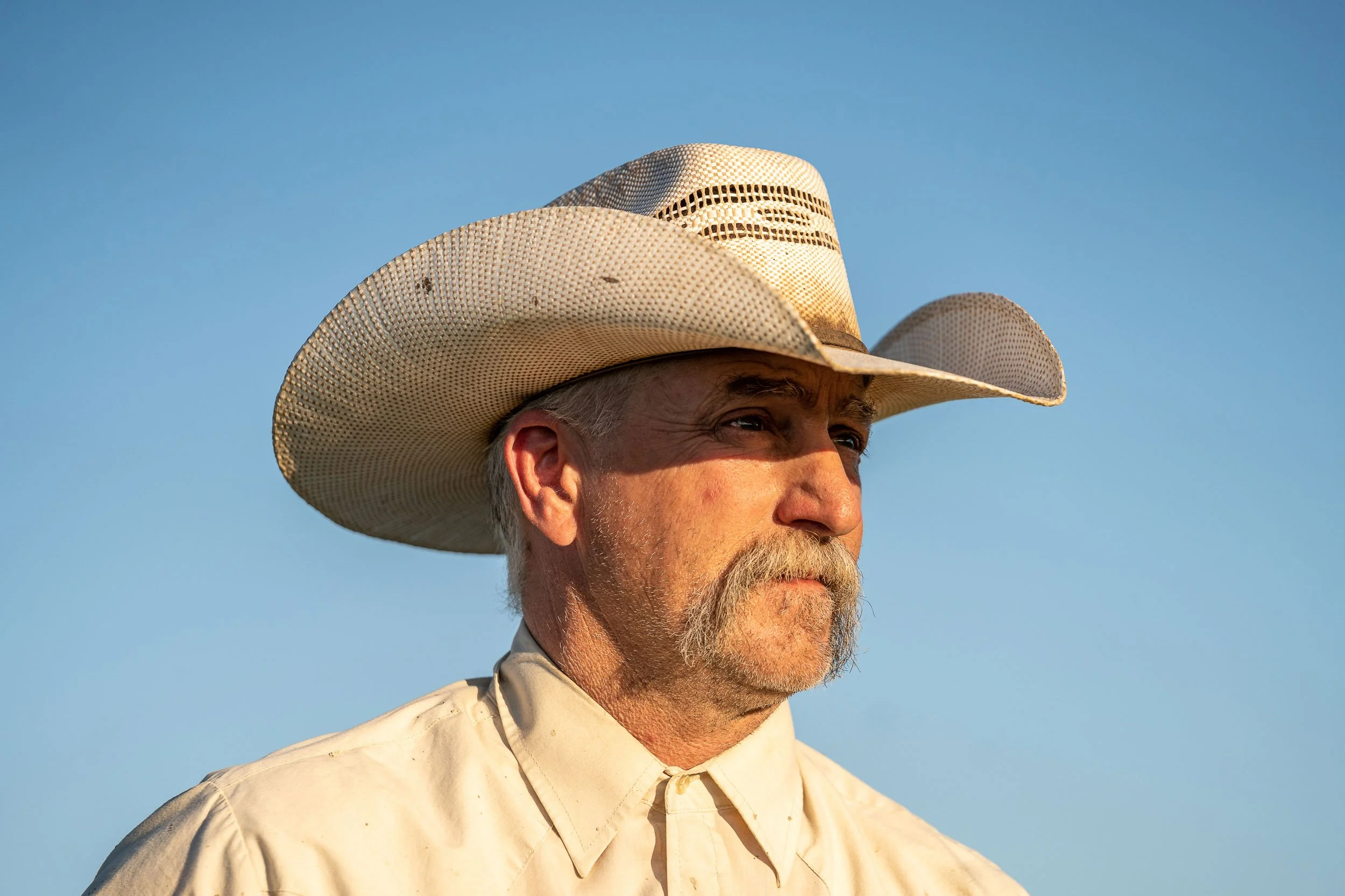 Close-up of an older man with gray hair, a beard, and mustache wearing a wide-brimmed straw hat and a beige shirt against a clear blue sky.
