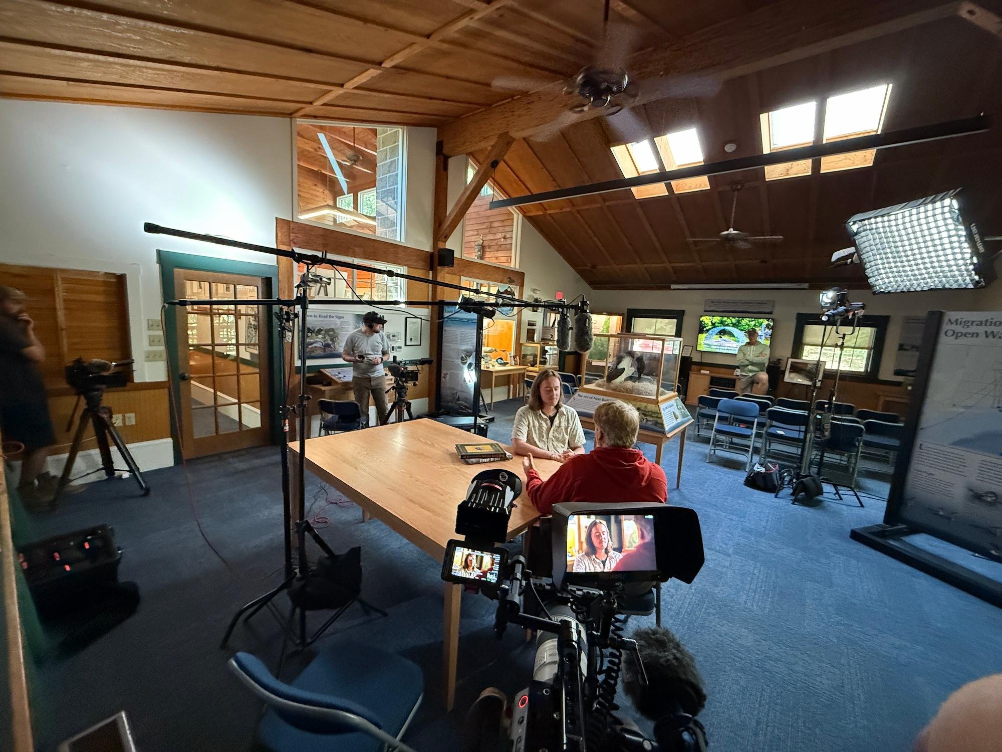 Inside a room with wooden ceiling, a TV screen and cameras set up filming two women at a table, with other people and display cases in the background.