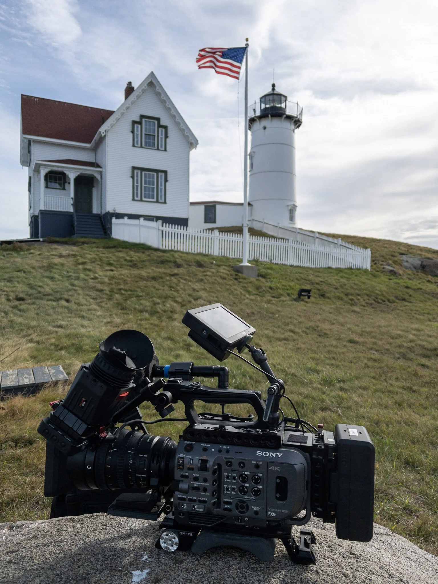 A professional camera setup with a camera on a tripod in the foreground, with a lighthouse and a house with a red roof in the background, atop a grassy hill with an American flag flying nearby, under a partly cloudy sky.