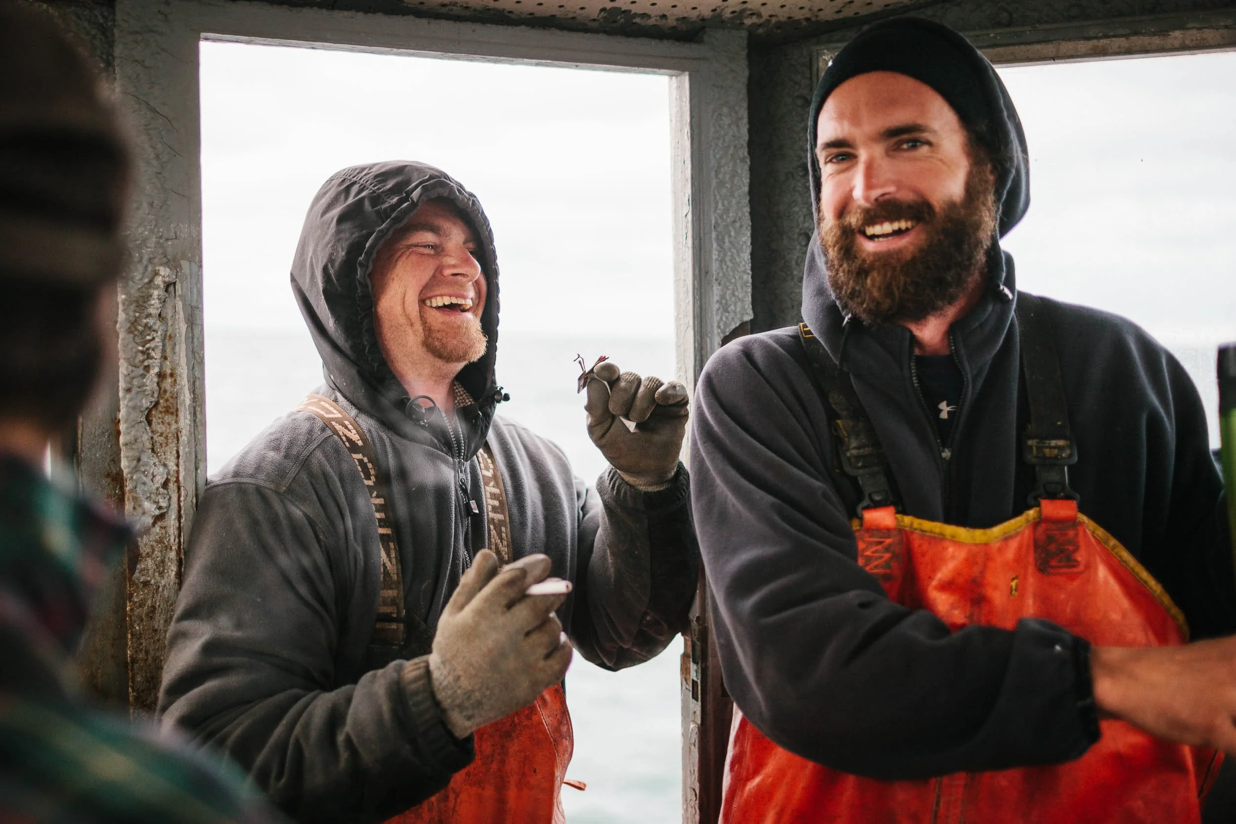 Two men wearing black beanies and outdoor jackets, smiling and talking inside a weathered wooden structure with open windows, overlooking a cloudy sky and water.