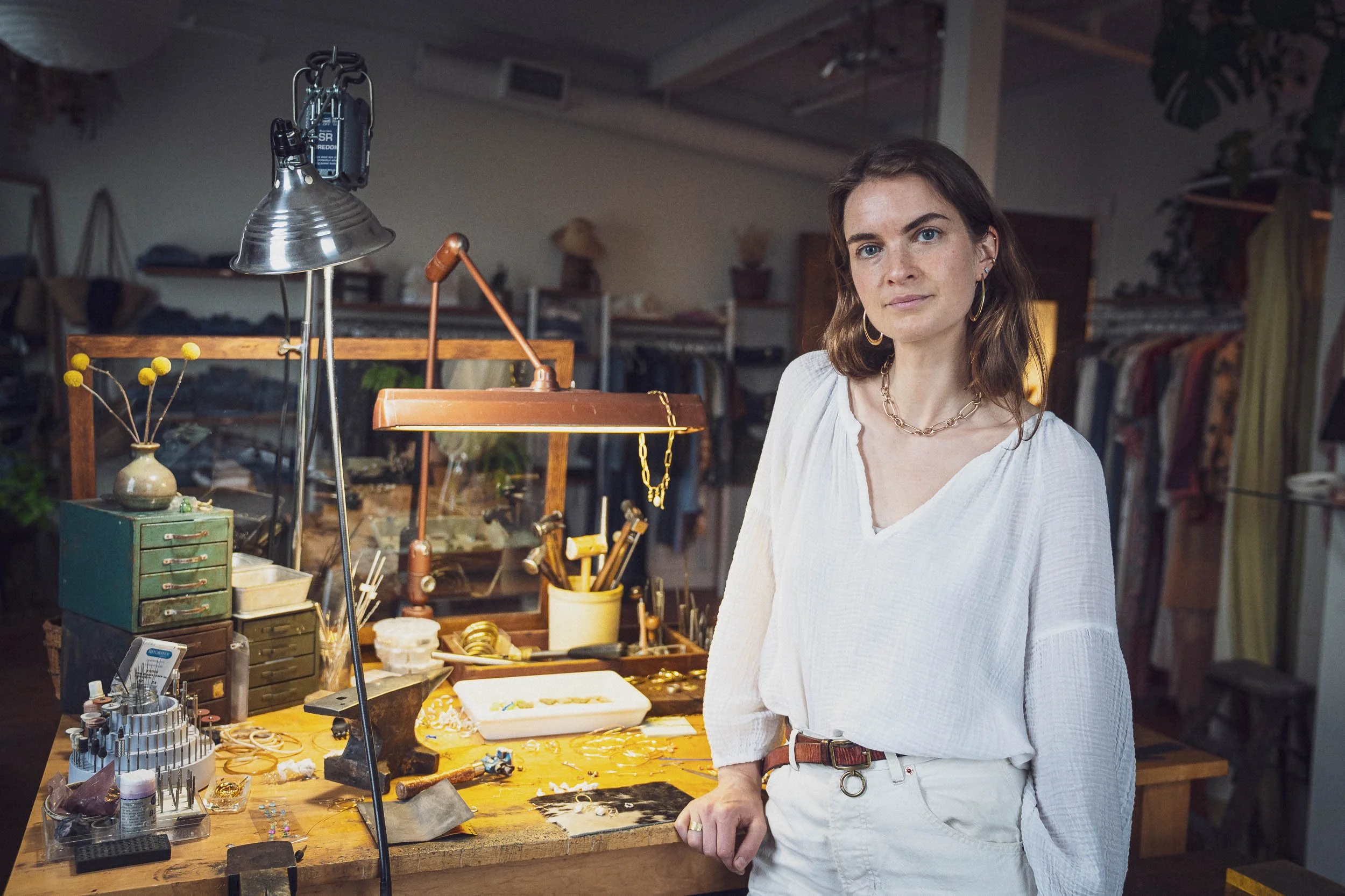 A woman with brown hair and gold hoop earrings stands at a jewelry workbench in a workshop, surrounded by tools, jewelry, and storage drawers.