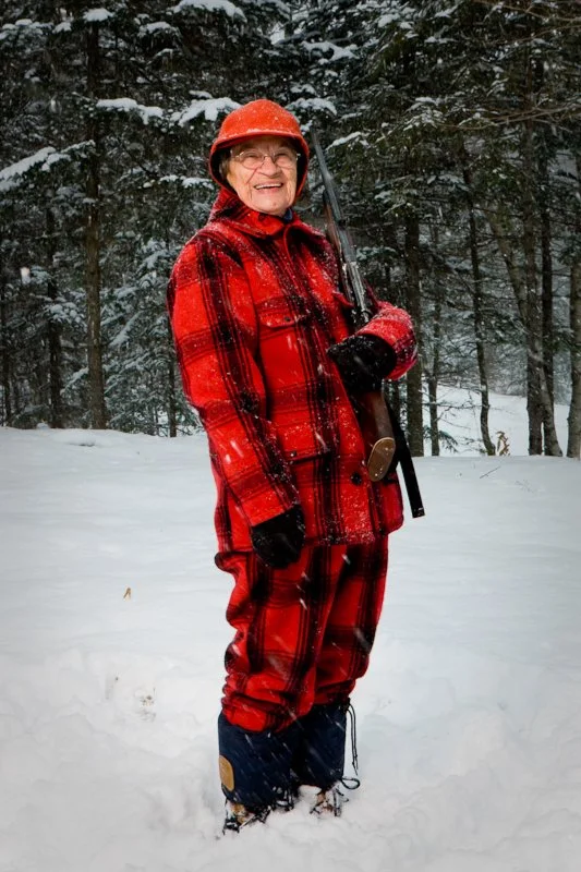 An elderly woman hunter in red plaid winter clothing stands in a snowy forest holding a rifle.