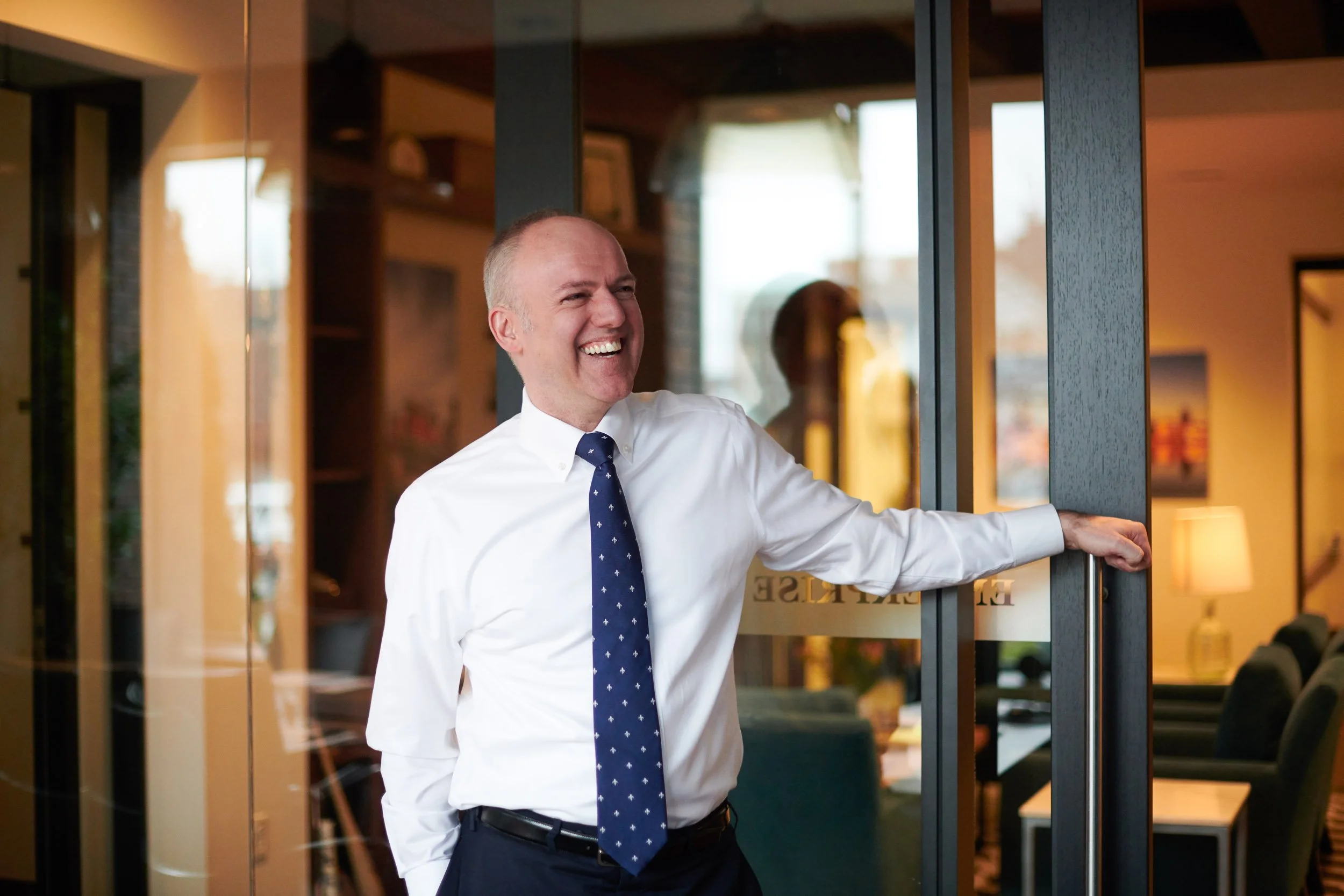 A man in a white shirt and navy blue tie smiling and opening a glass door to a restaurant or cafe, with warm lighting and a cozy interior visible inside.