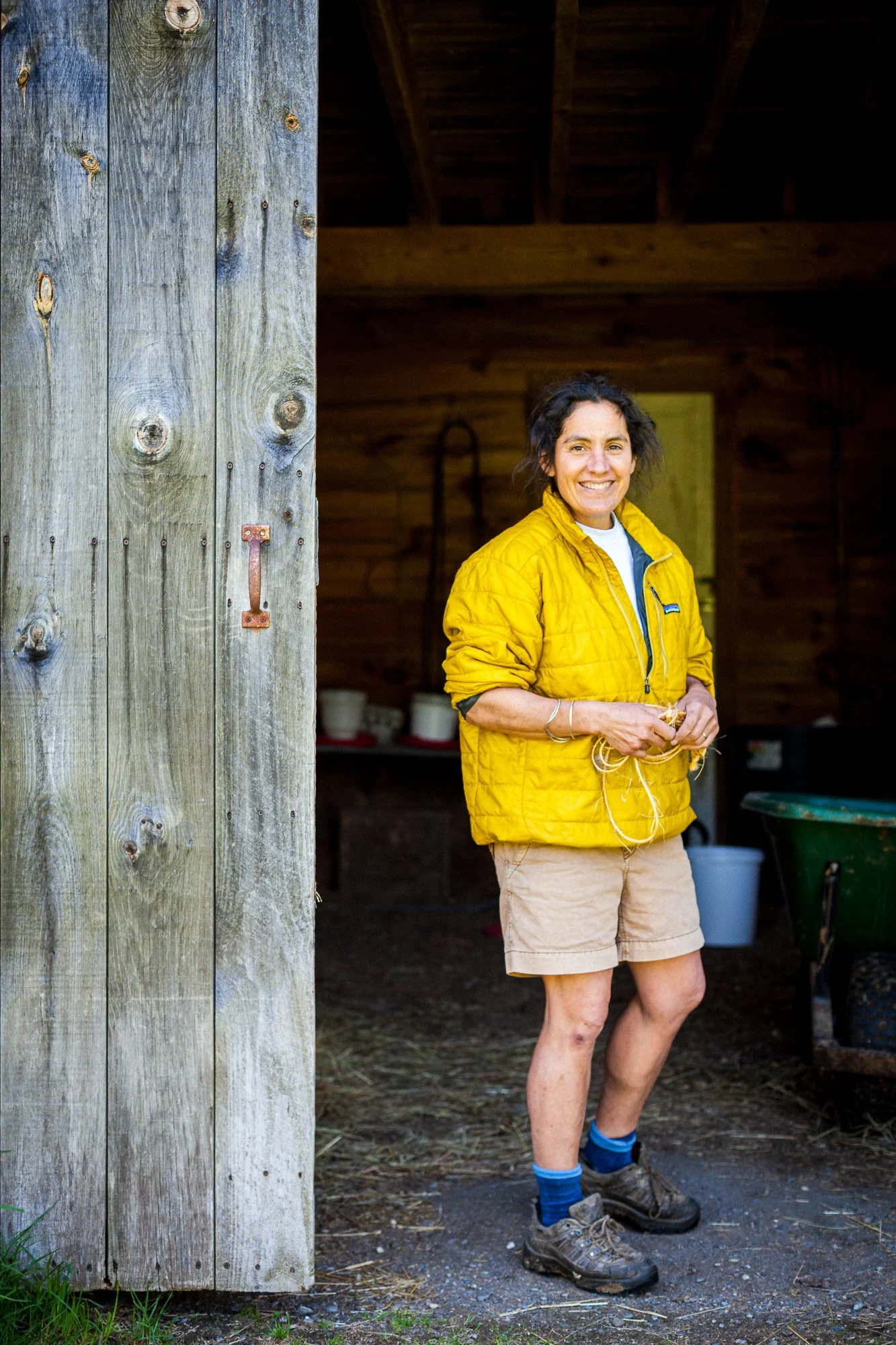 A woman farmer stands partially inside a wooden shed, smiling at the camera, wearing a yellow jacket, beige shorts, and hiking boots with blue socks. The shed has a large wooden door and visible interior with some buckets and gardening tools.