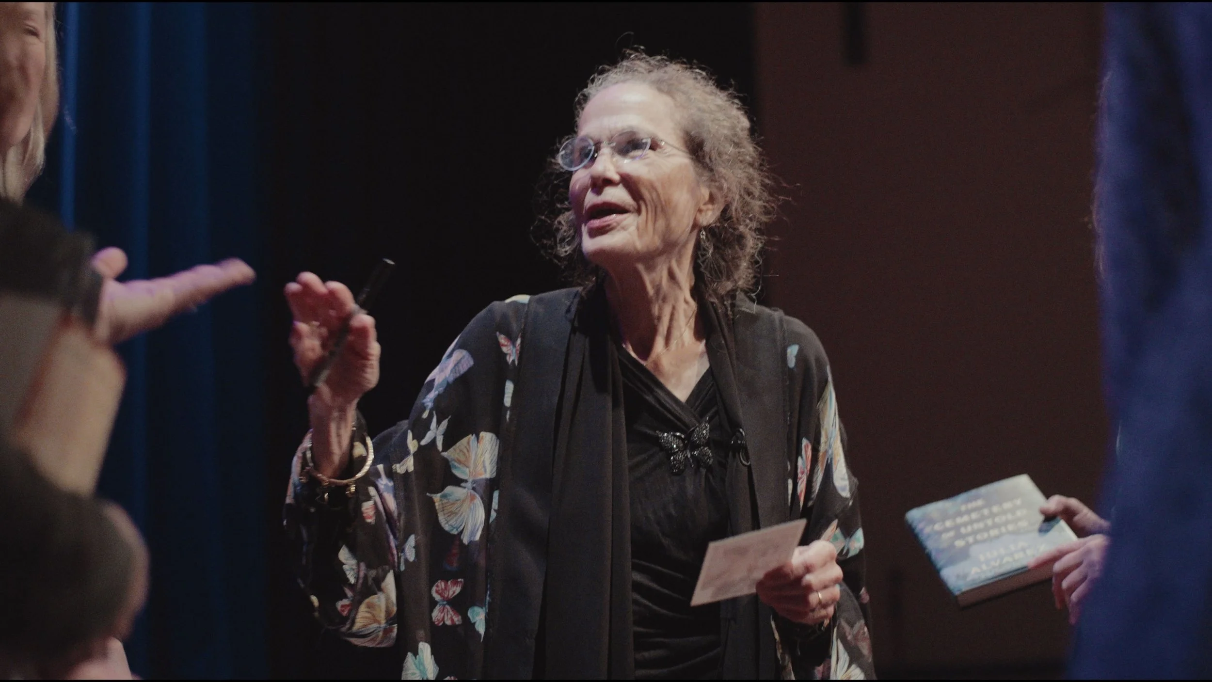 Julia Alvarez wearing glasses, a butterfly brooch, and a dark floral jacket talking to a group of people, holding a small piece of paper, with another person holding a book for the PBS show American Masters.