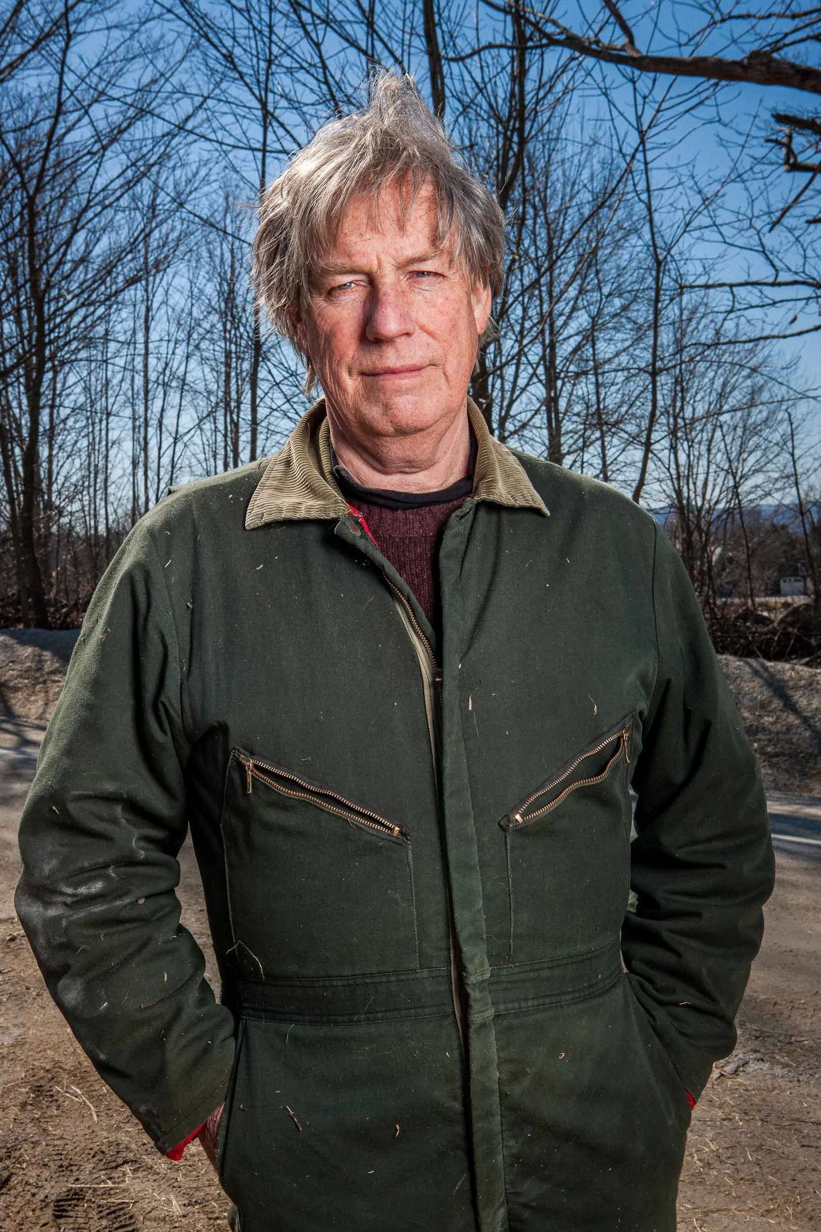 A man with gray hair and a serious expression, wearing a green jacket, stands outdoors in front of bare trees during daytime.