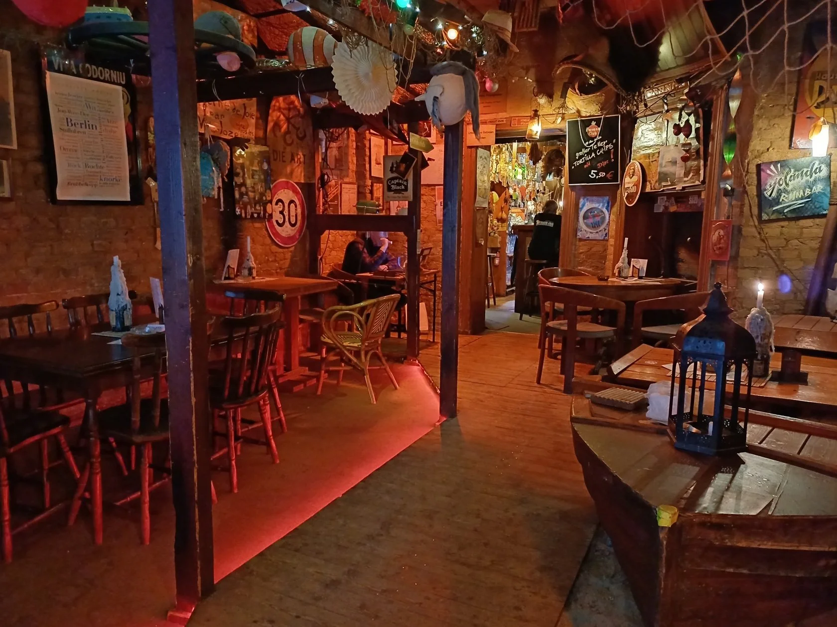 Innenansicht eines gemütlichen Restaurants oder einer Bar mit rustikaler Dekoration, Holztischen und Stühlen, bunten Lichtern und verschiedenen Dekorationsstücken an den Wänden.