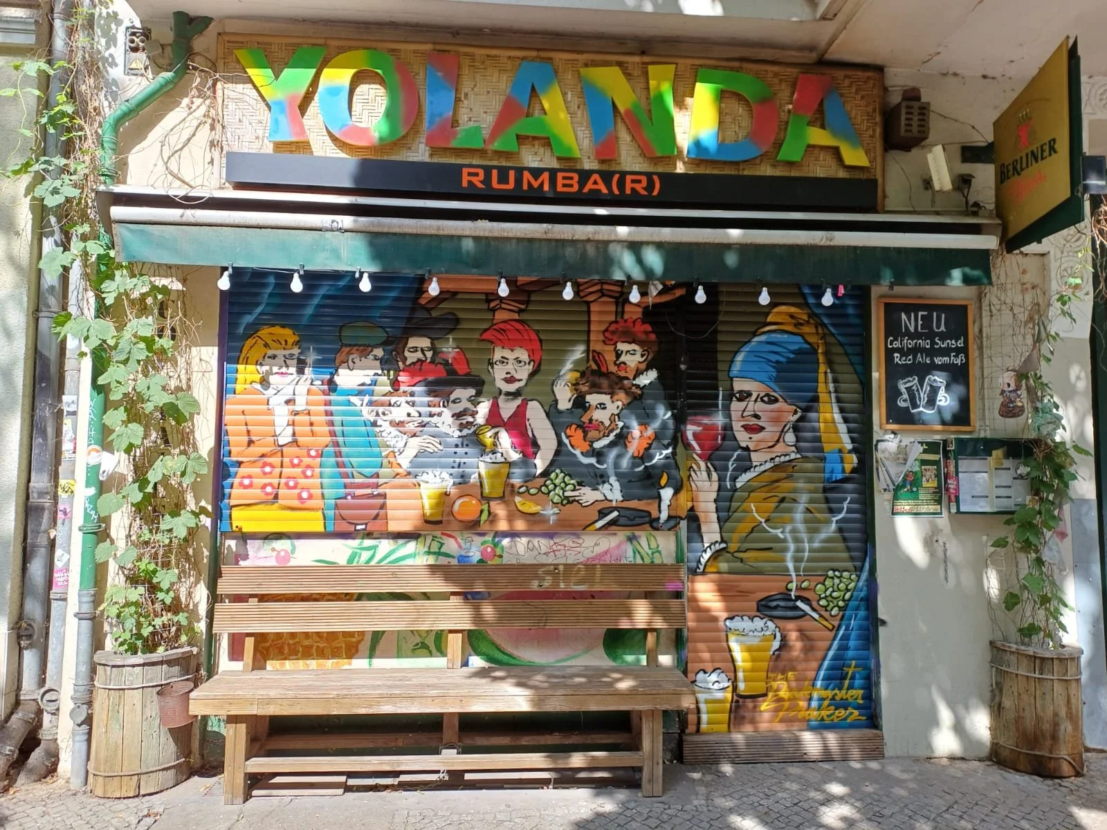 Straßenszene vor einem Laden namens "YOLANDA RUMBAR" mit bunter Graffiti-Dachzeichnung und lachenden, feiernden Personen, die Bier trinken, eine Zigarette rauchen und eine Party feiern.