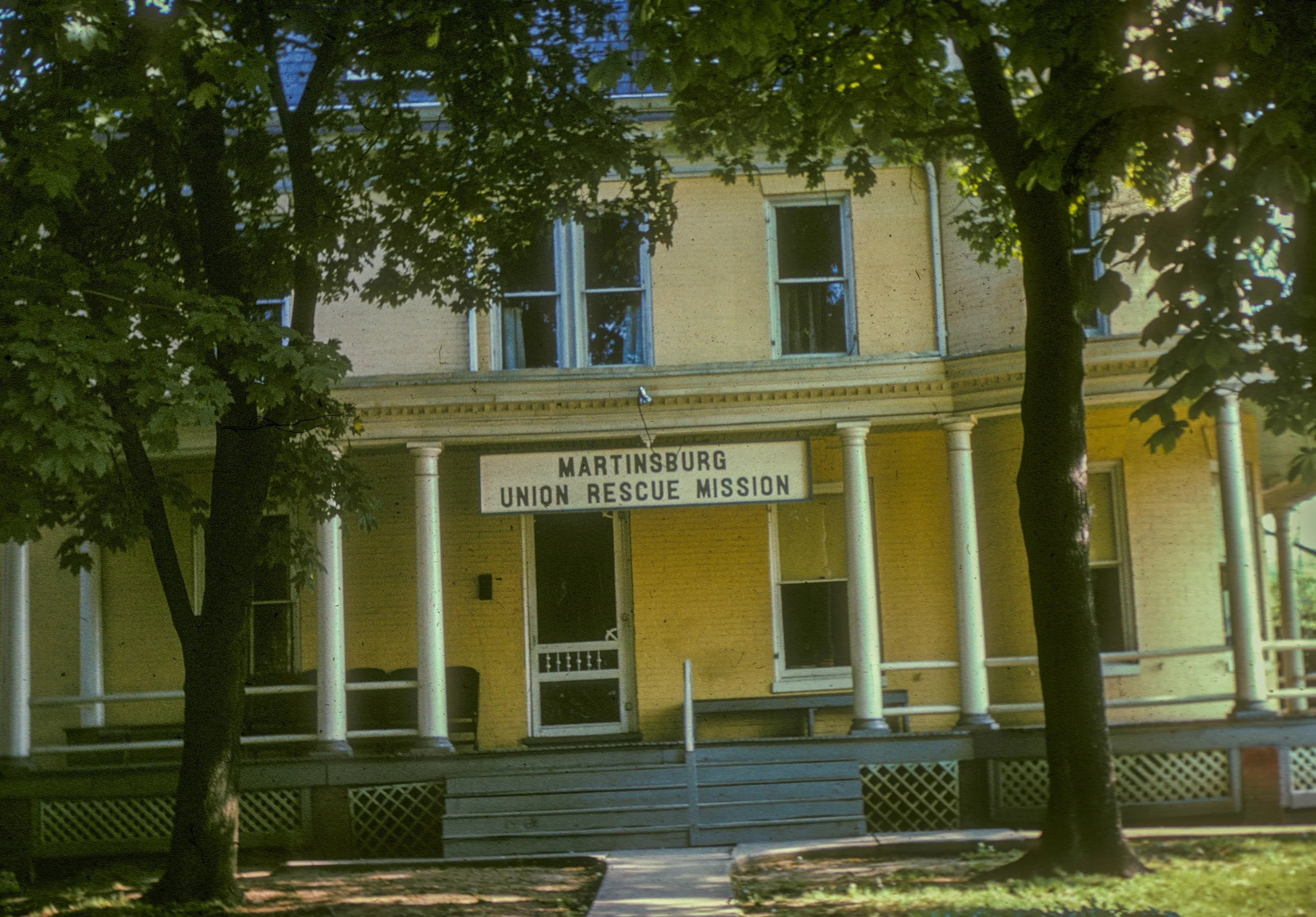 A yellow, two-story building with a porch, surrounded by trees. The sign on the building reads "Martinsburg Union Rescue Mission."