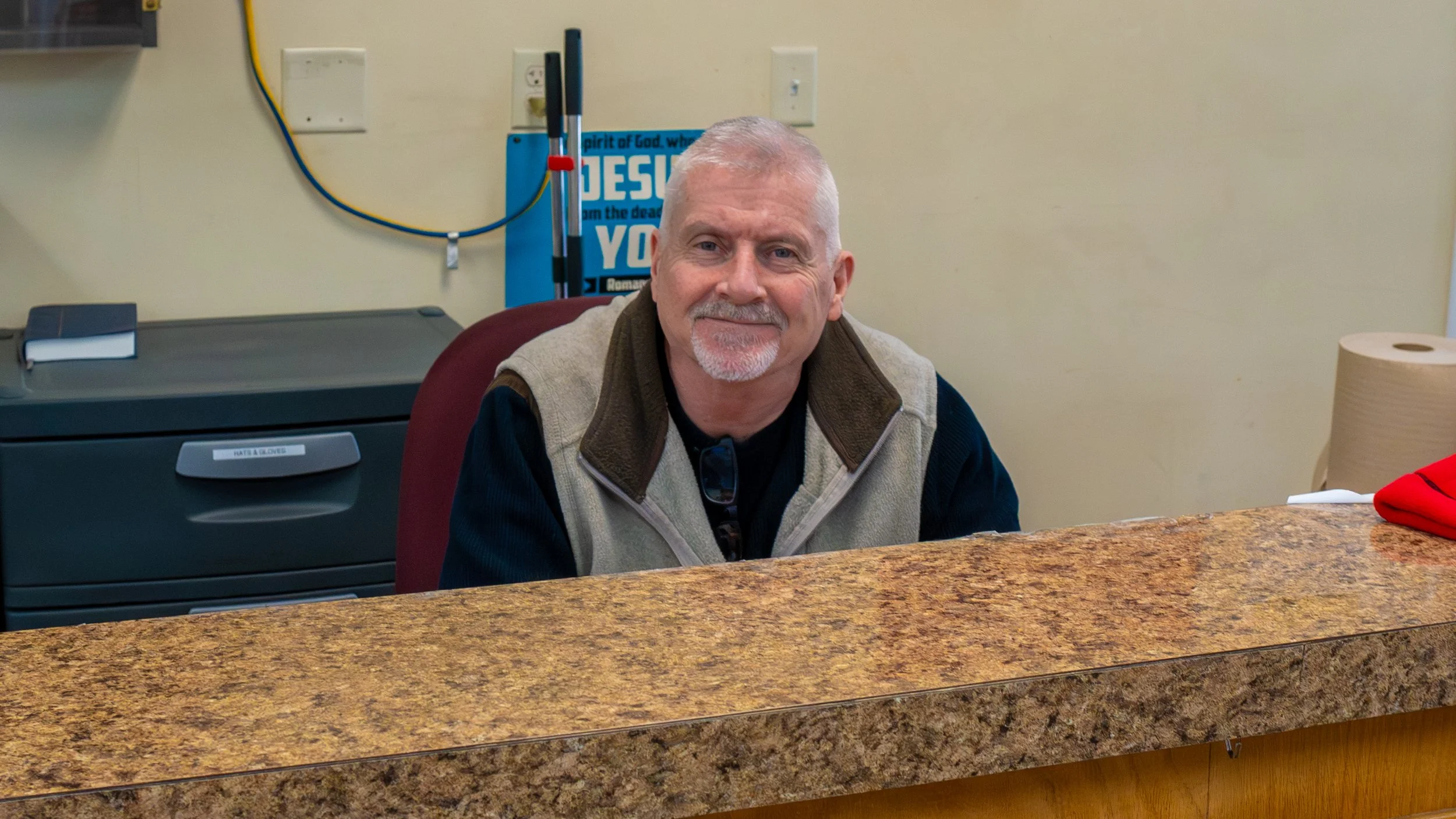 A middle-aged man with gray hair and a beard sitting behind a marble countertop, smiling at the camera, wearing a beige and dark jacket. In the background, there are electrical outlets, a blue sign with religious text, a trash can, and cleaning tools.
