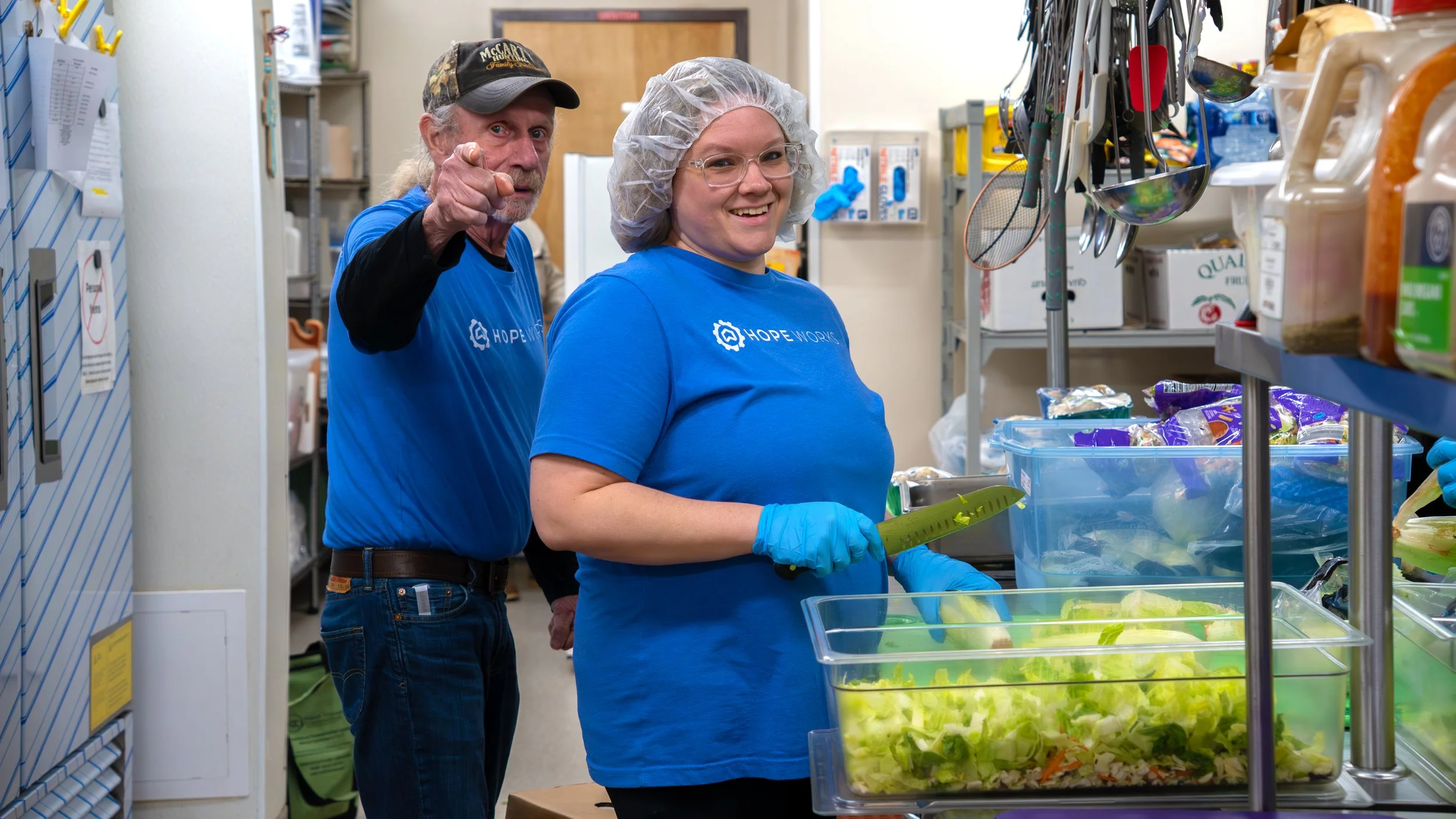 Two volunteers wearing blue HopeWorks T-shirts, gloves, and hairnets in a food pantry, one woman holds a knife and the man points towards the camera.