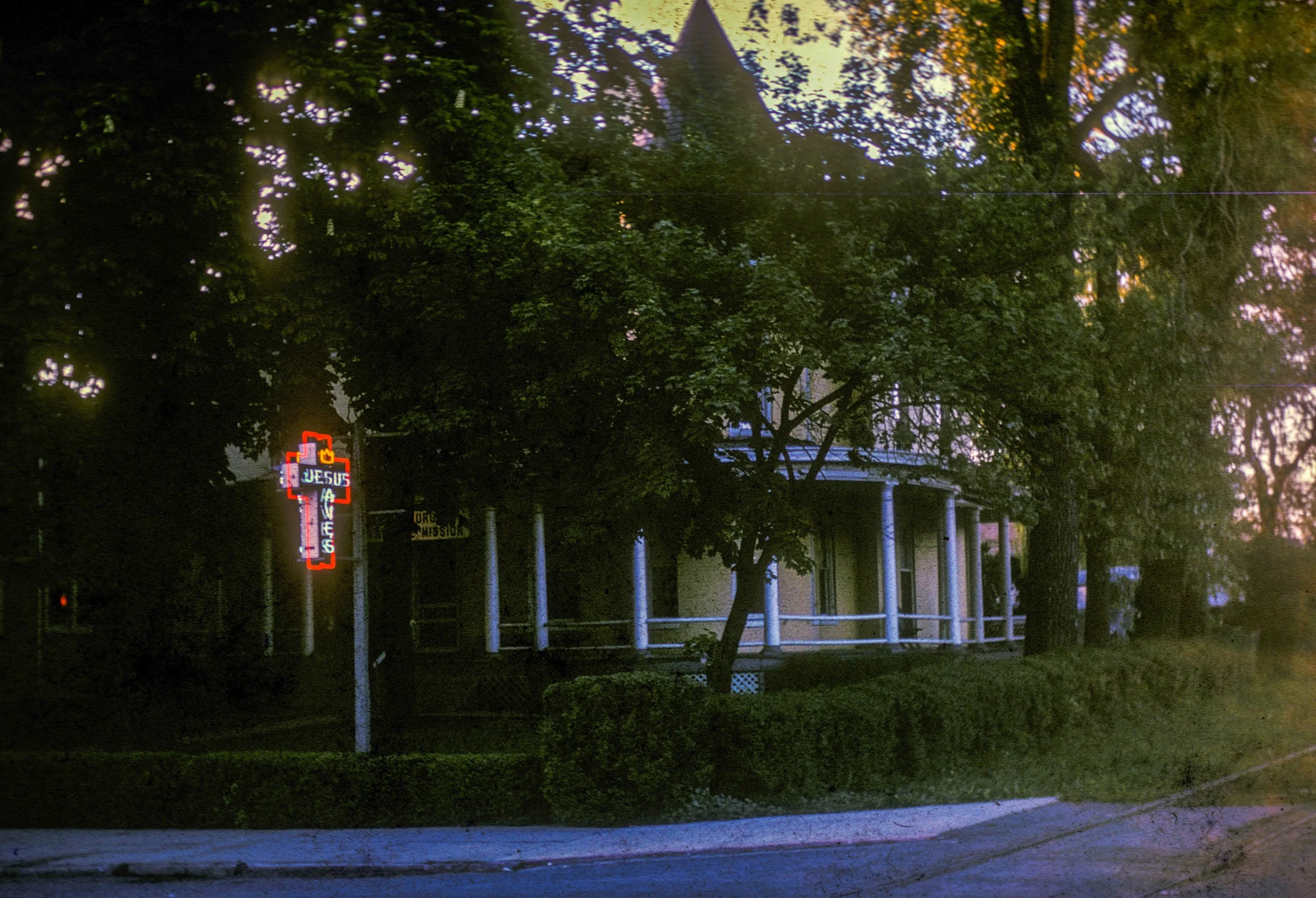 A house partially obscured by large trees with a neon sign that reads 'JESUS LOVES' in red, white, and blue lights. The house has a porch with white railings and is set along a quiet street in twilight.