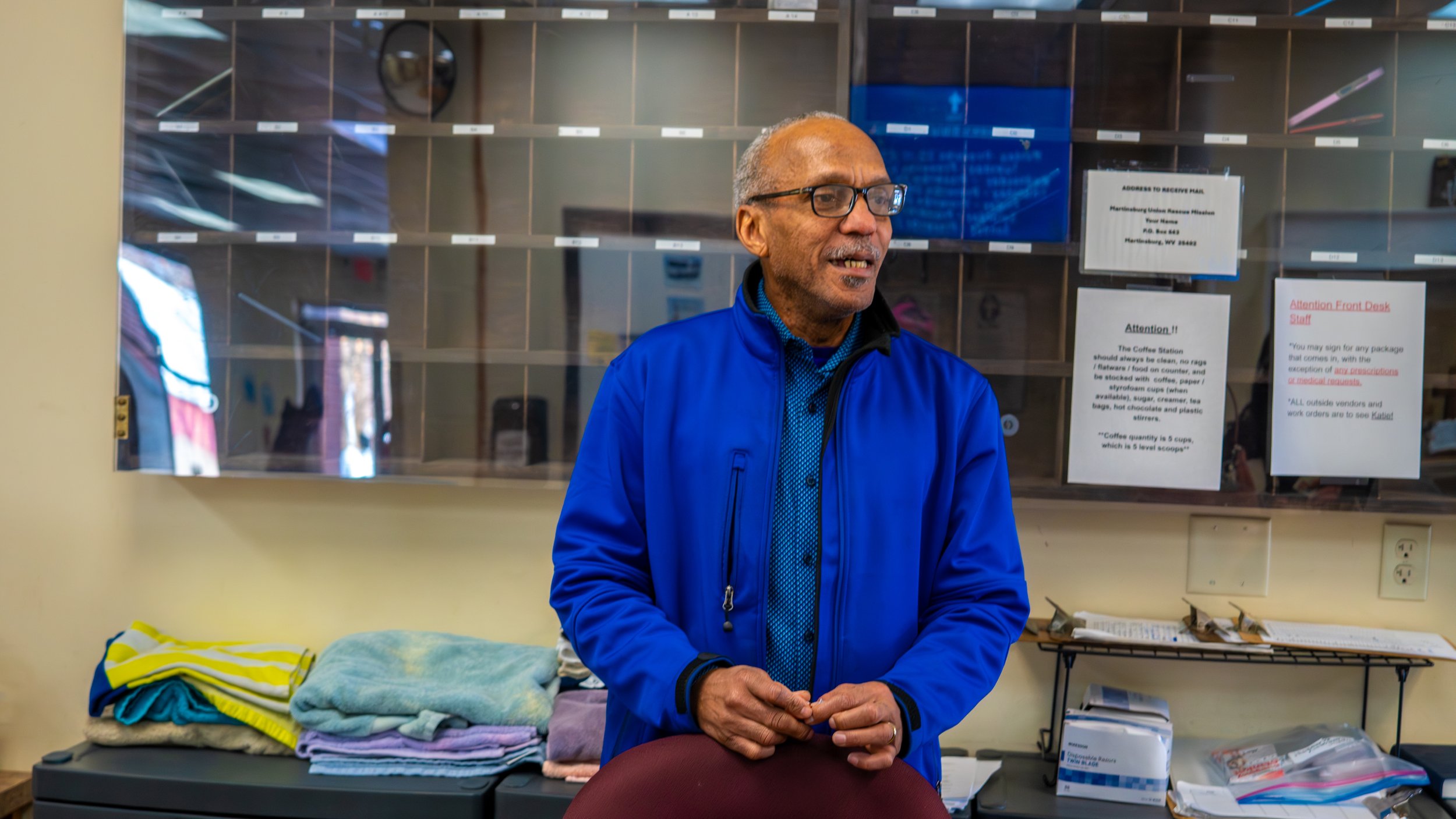 An older man in a blue jacket and glasses stands behind a counter in an office or service area, with linens and papers on the counter and notices on the wall behind him.