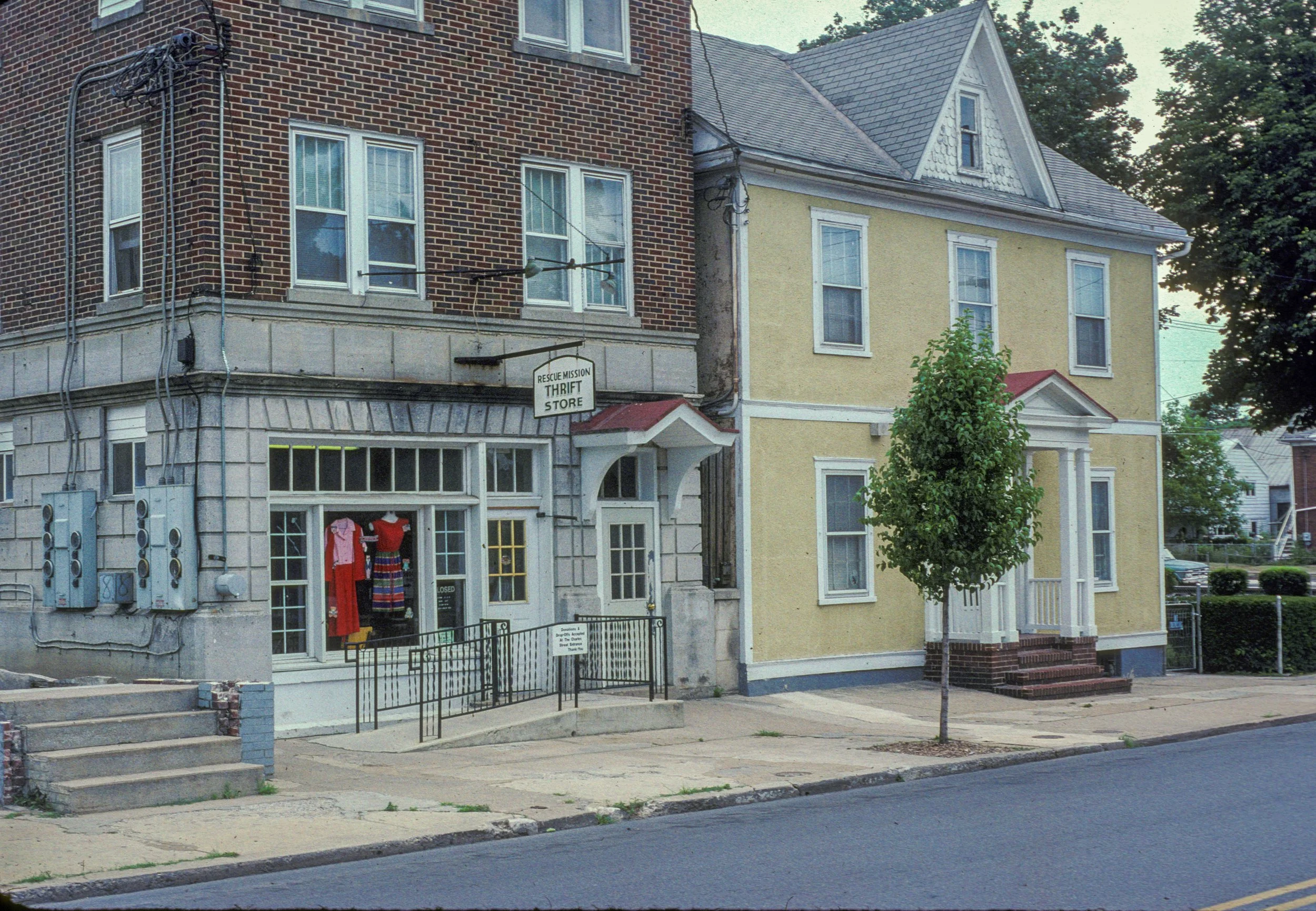 Street view featuring a thrift store with mannequins wearing colorful dresses in the window, adjacent to a yellow residential building with steps leading to the entrance, small trees lining the sidewalk, utility boxes and pipes on the building's wall, and a calm street under a clear sky.