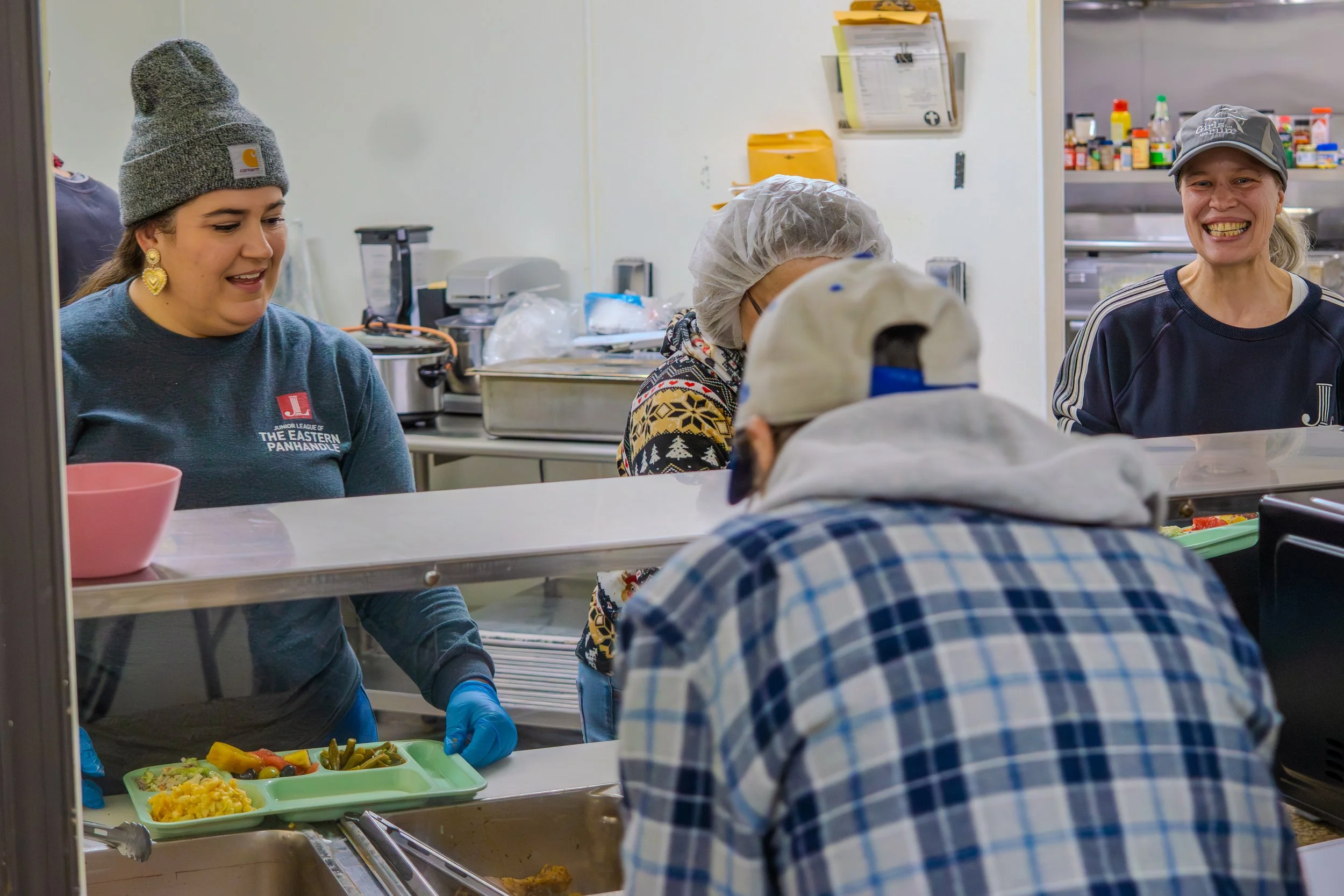 Four women and one man working in a kitchen, serving food to an elderly man. The women are smiling and wearing casual clothes, with two of them wearing hats and one wearing a hairnet. The elderly man has a cap and a plaid shirt. The kitchen has various equipment and supplies in the background.