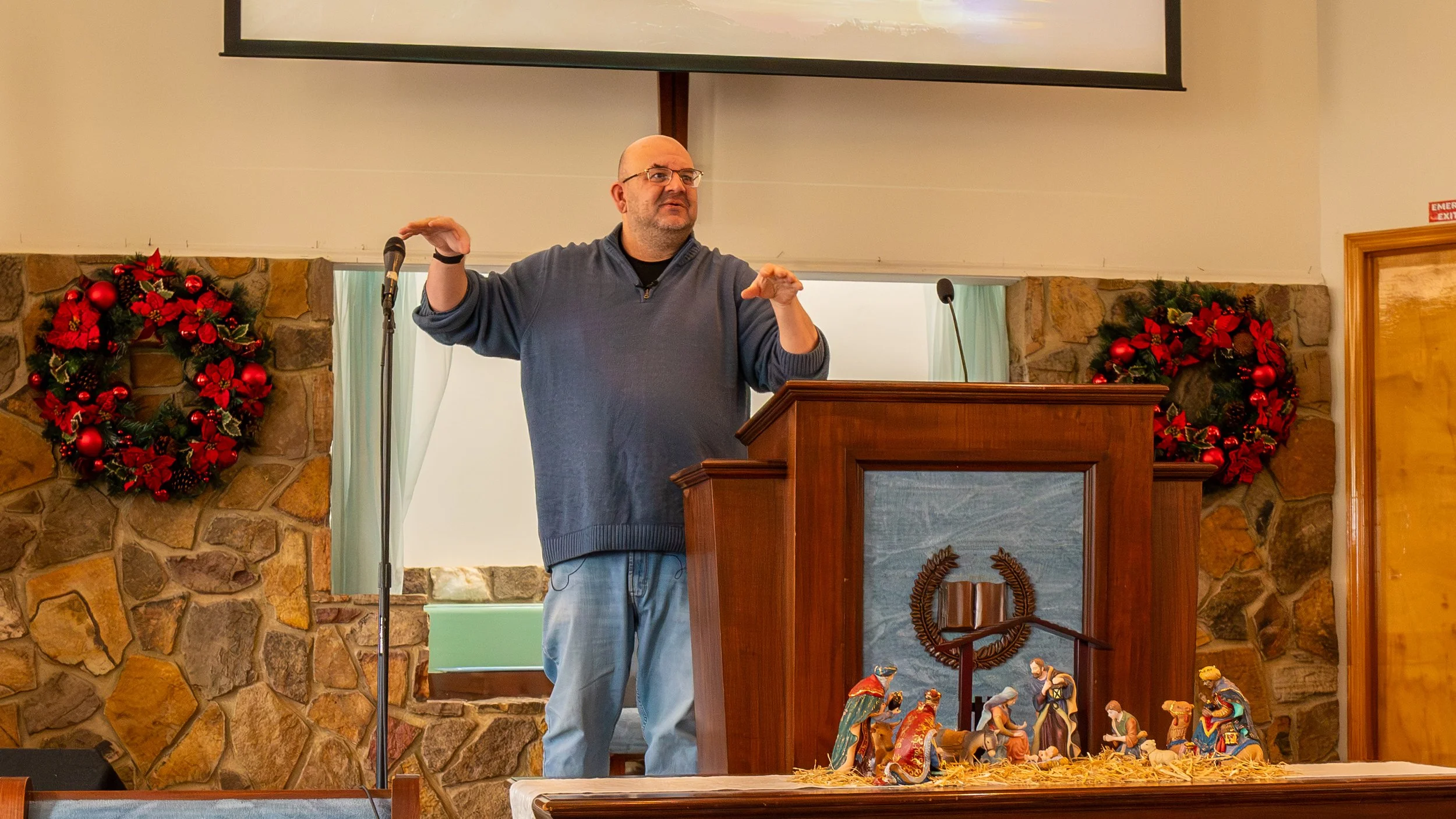 A man standing behind a wooden lectern decorated with nativity figurines, gesturing with his hands, with Christmas wreaths on stone walls behind him.