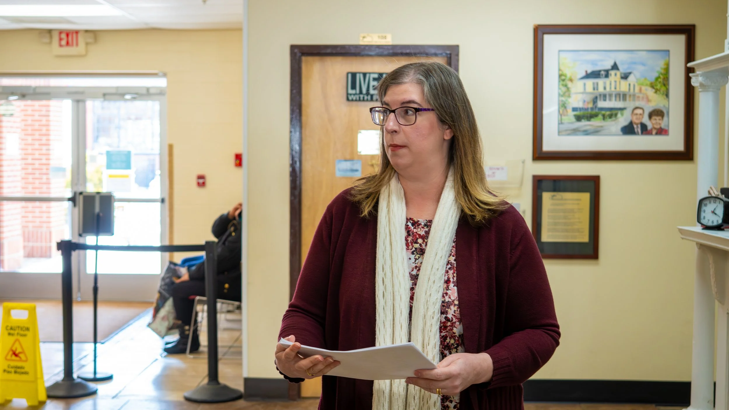 A woman with glasses, wearing a burgundy cardigan and cream-colored scarf, holding papers inside a building with framed pictures on the wall and a waiting area in the background.