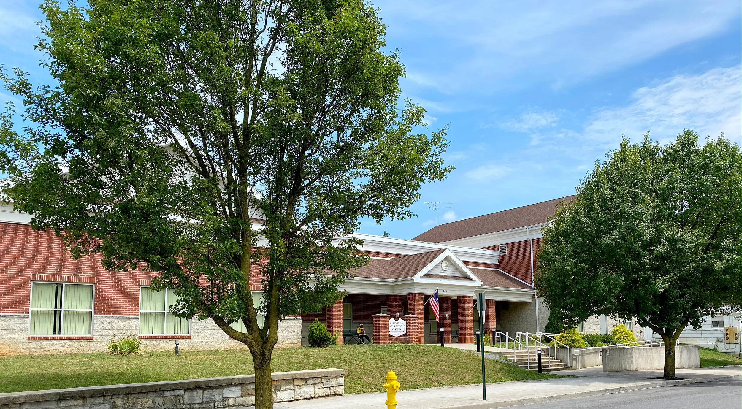 A brick building with a brown roof and a white entrance porch, two trees in front, a yellow fire hydrant on the sidewalk, and a person sitting on a bench near the entrance, under a partly cloudy sky.