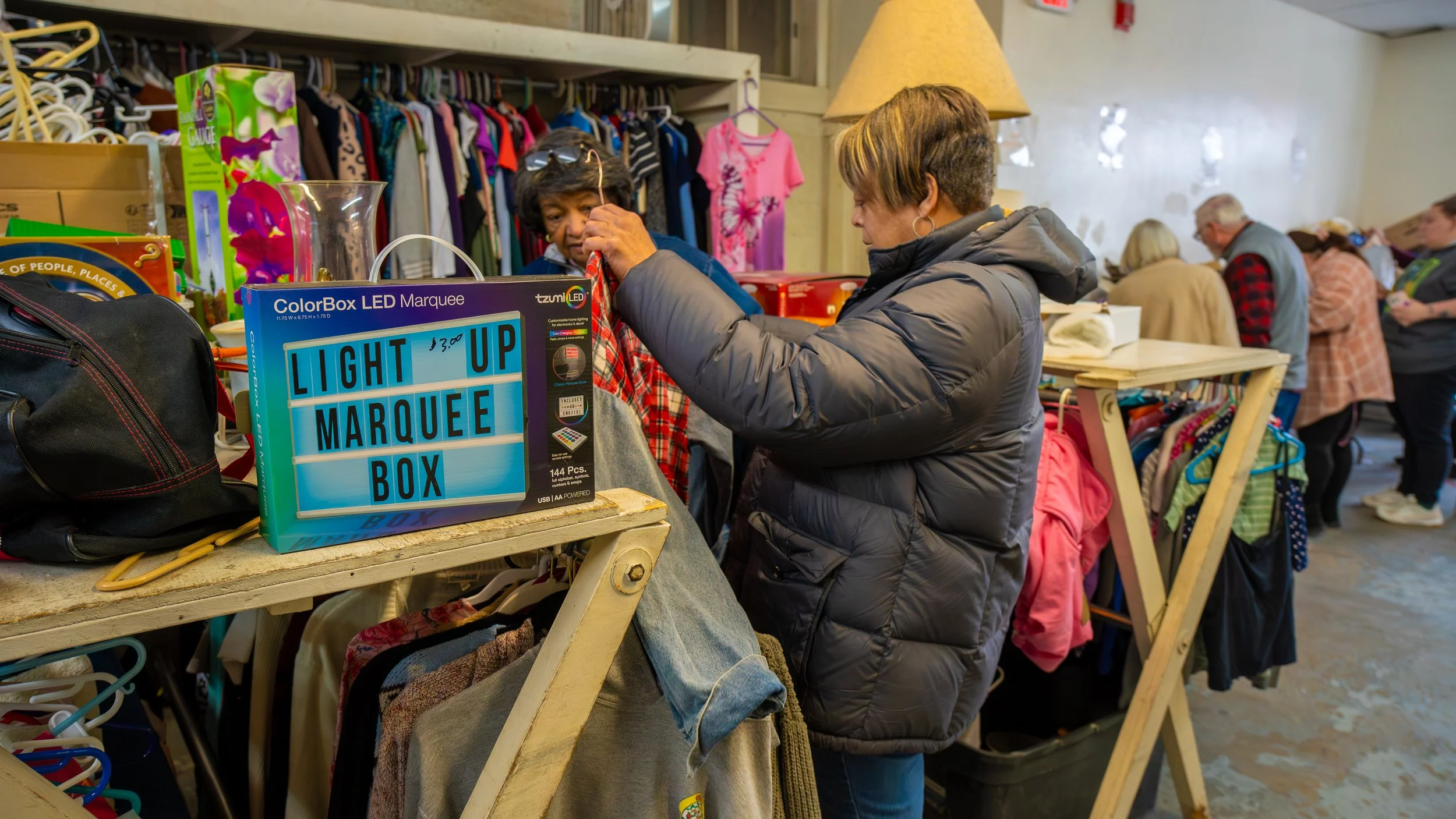 A woman shopping at a thrift store or flea market, browsing clothing racks and items on tables, with other shoppers in the background.