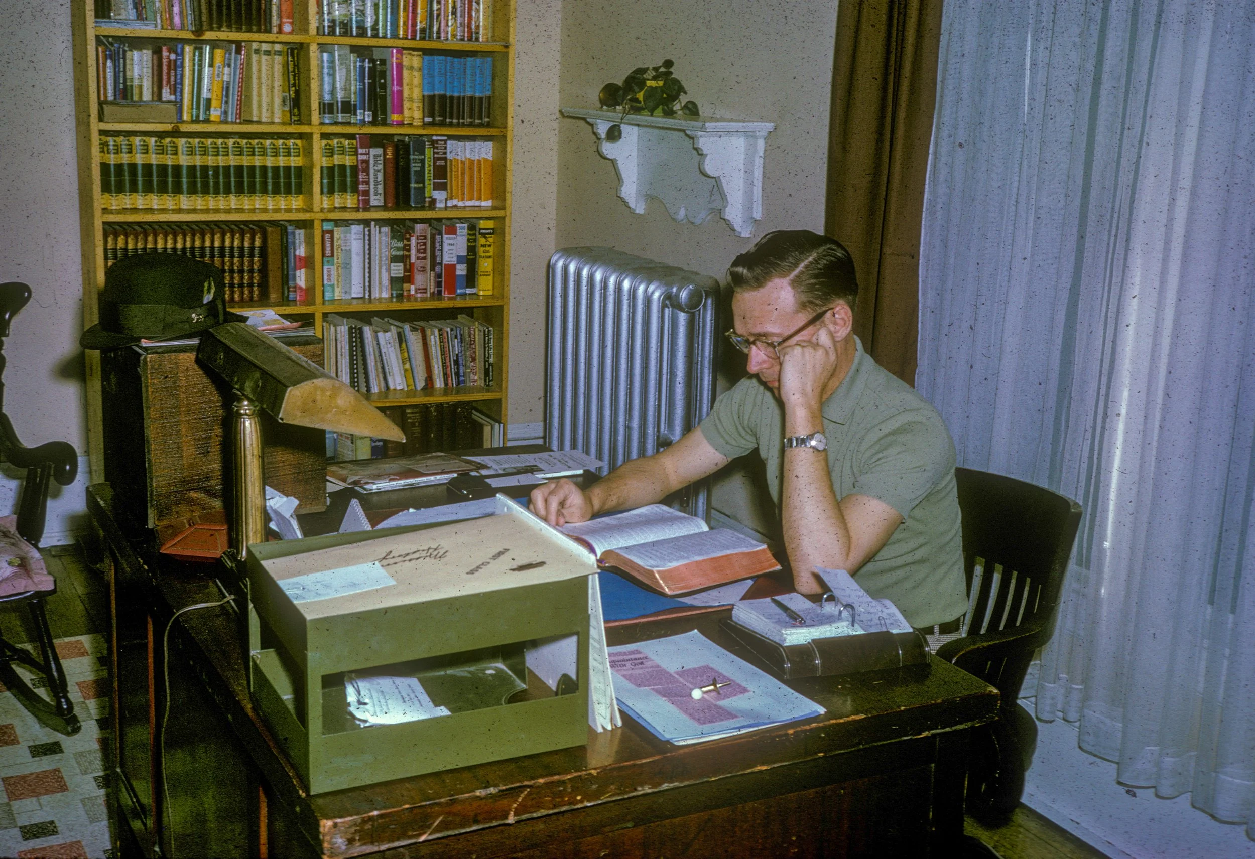 A man with glasses, wearing a green shirt, sitting at a wooden desk with books, papers, and a typewriter, reading a book in a home office with a bookshelf, radiator, and curtains.