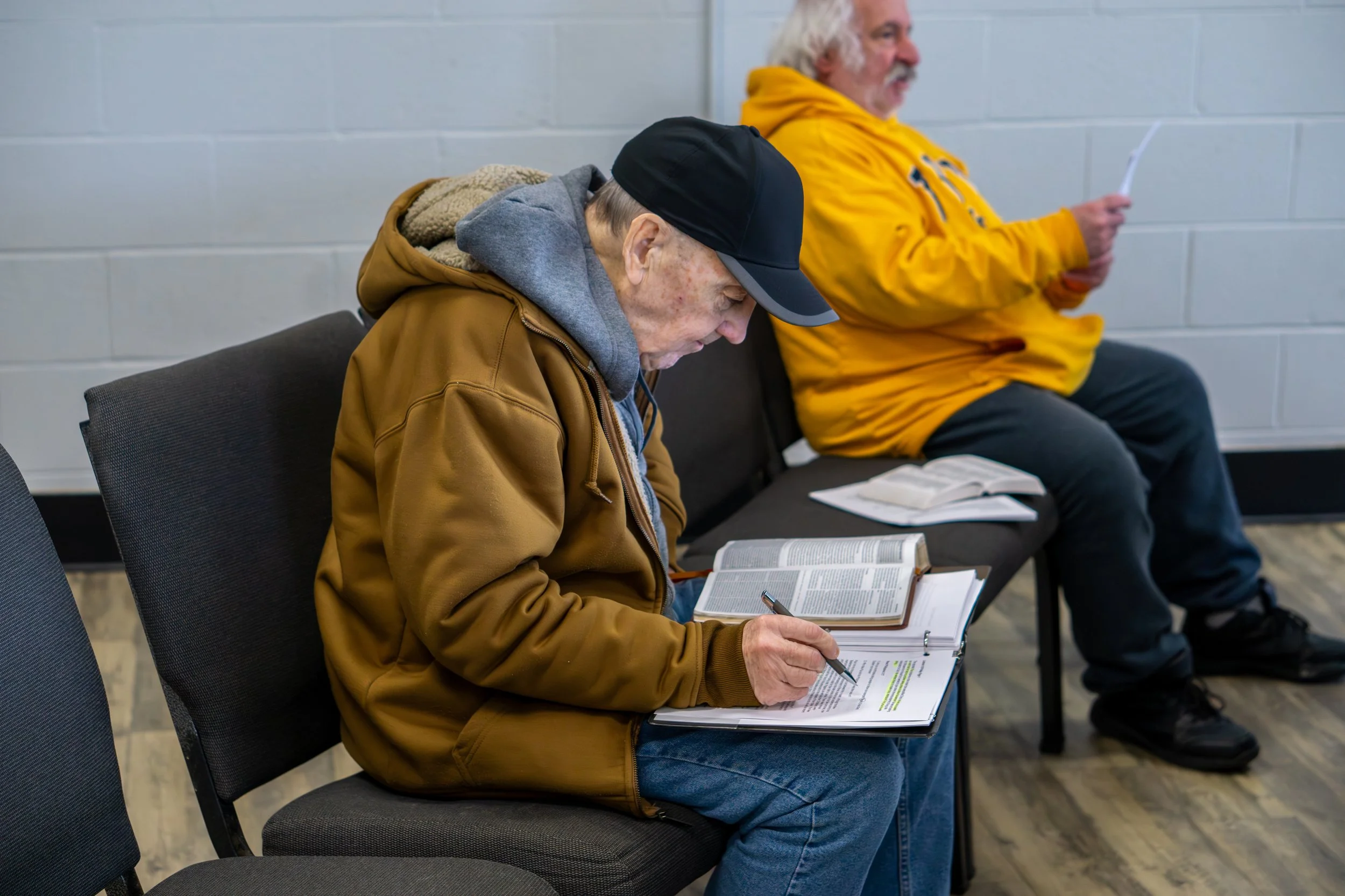 Two men sitting on a black bench in a waiting room, reading and taking notes from open Bibles.