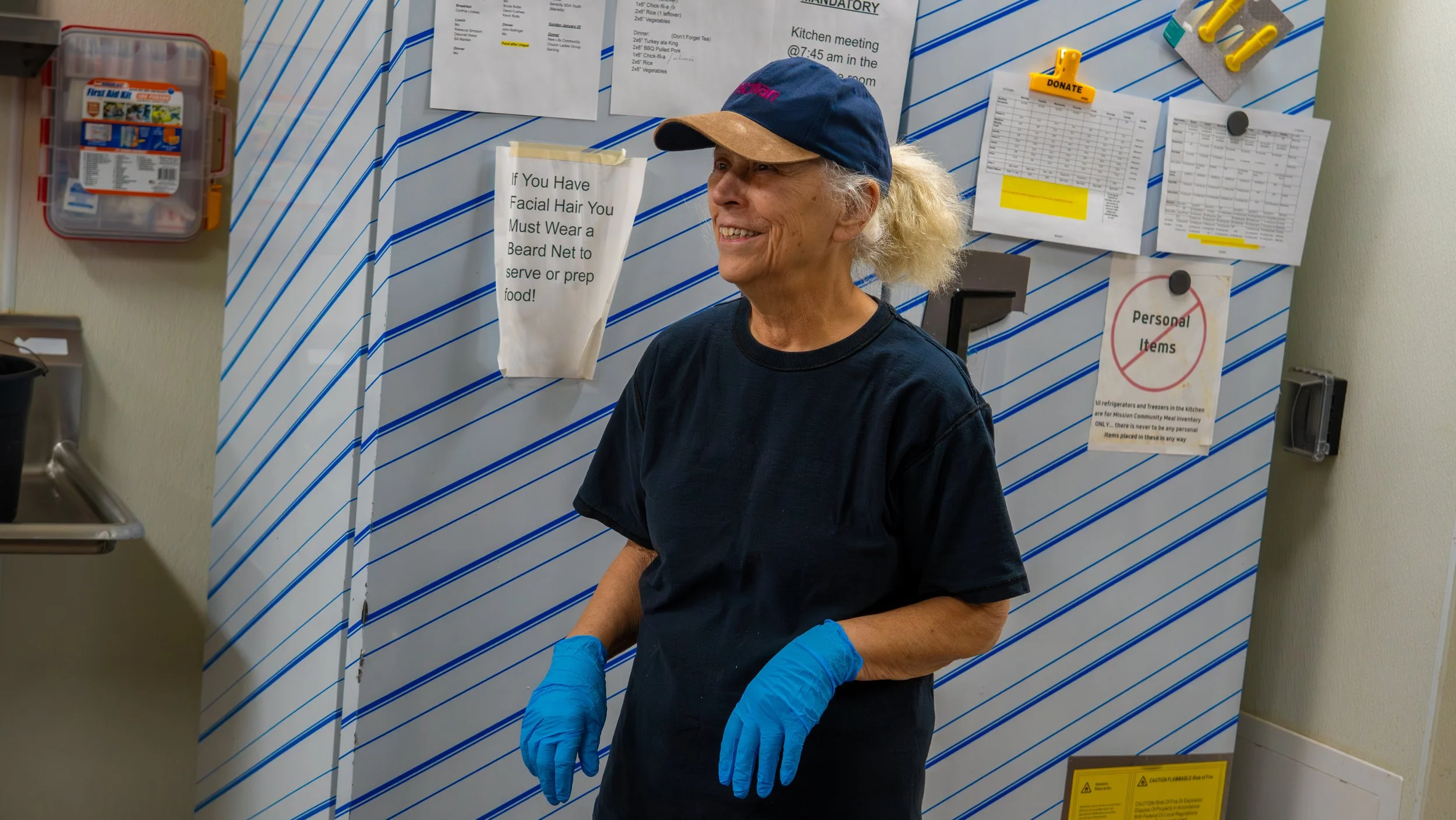A smiling woman with gray hair tied in a ponytail, wearing a navy baseball cap, black t-shirt, and blue gloves, stands in front of a wall with notices and papers pinned on it.