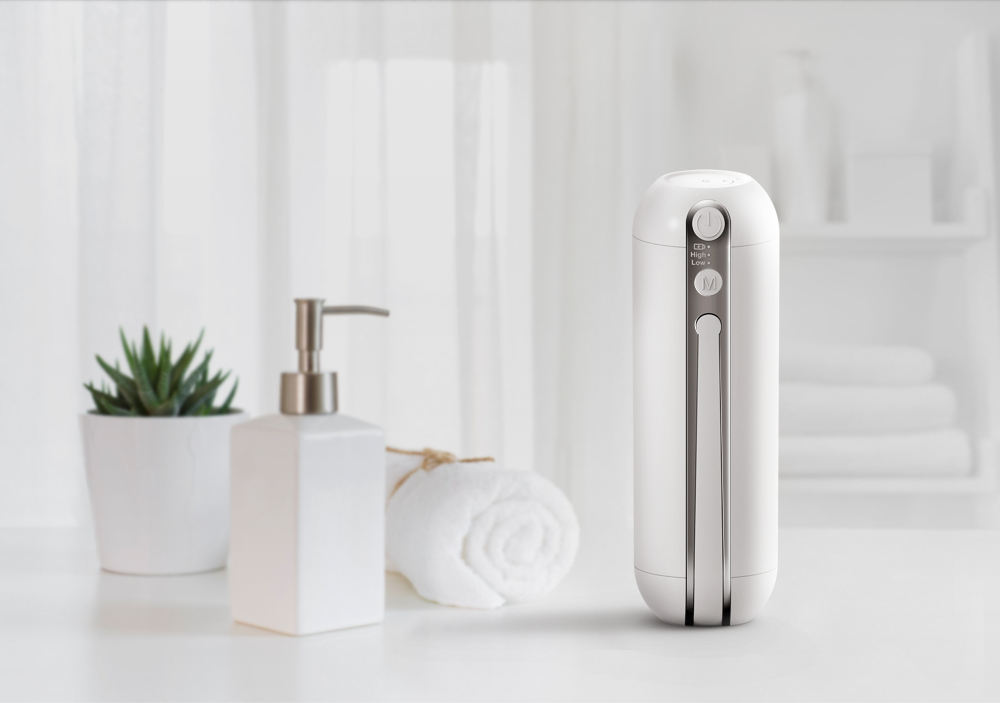 A white portable bidet with a digital control panel on top, placed on a white bathroom countertop next to a potted succulent plant, a white rolled towel, and a white soap dispenser.