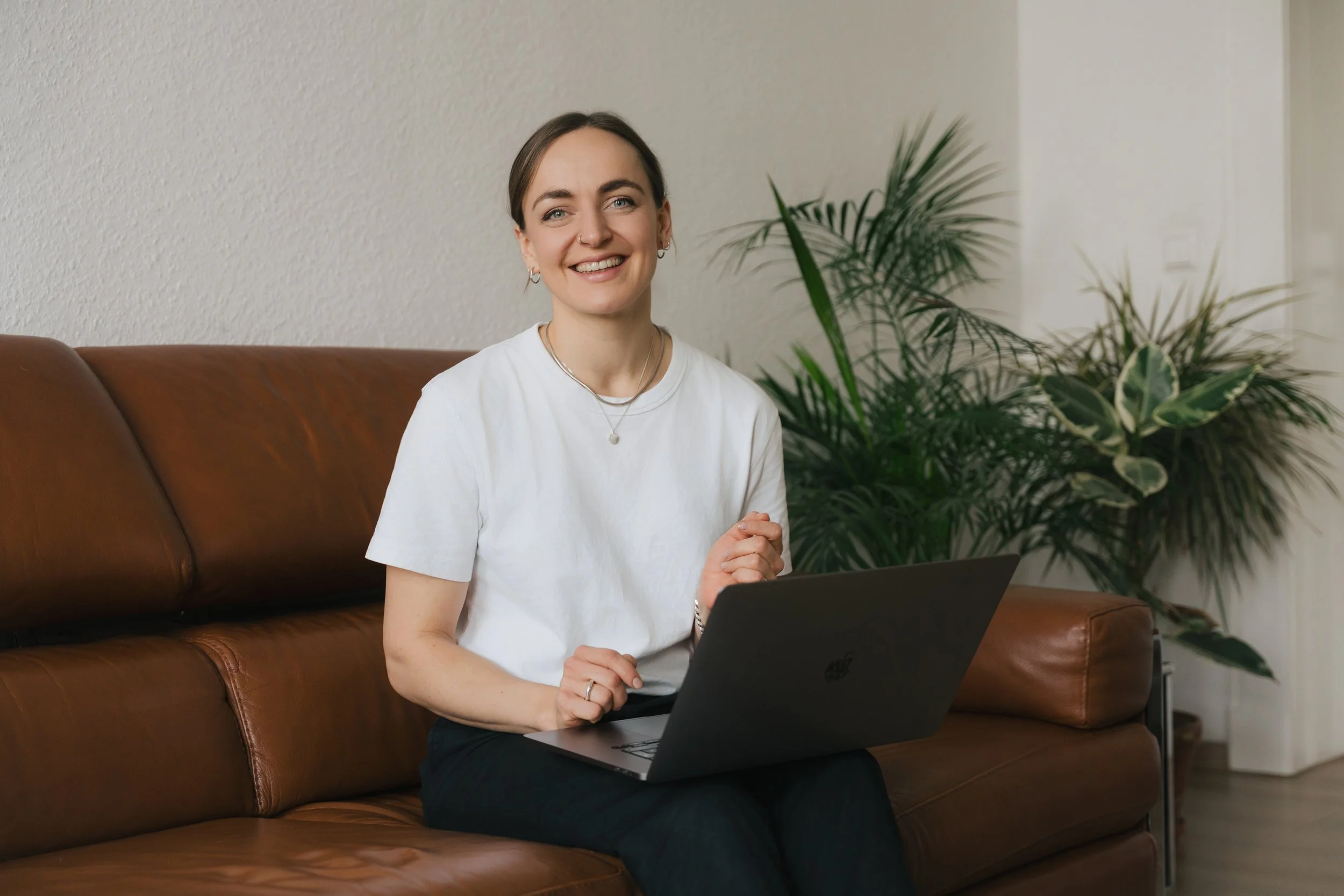 Antonia Grüner, Ordnungscoach, mit braunen Haaren, weißen T-Shirt und schwarzer Hose sitzt auf einem braunen Sofa vor einem offenen Laptop, umgeben von grünen Pflanzen, in einem modern eingerichteten Raum.