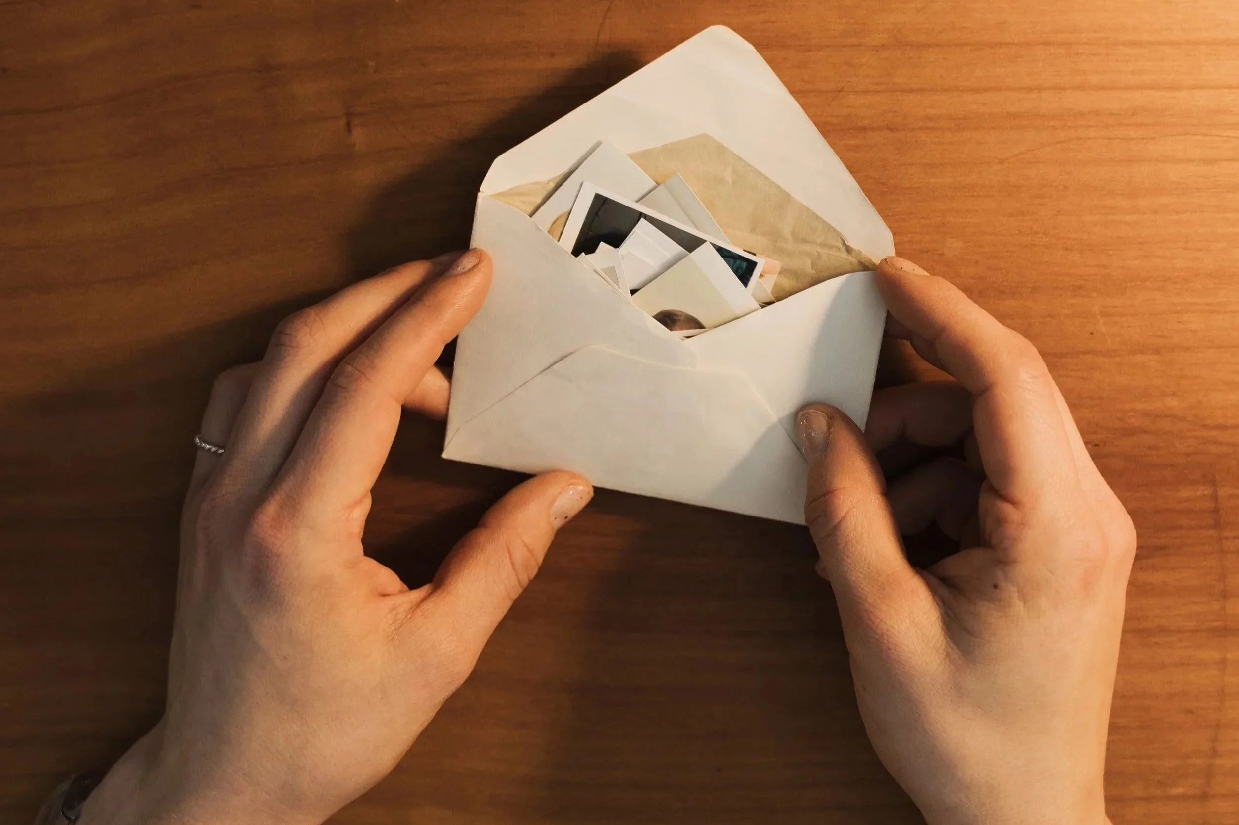 Hände, die einen Papierumschlag öffnen, auf einer Holztischplatte. Es sind die Ecken von Fotos in dem Umschlag zu sehen.