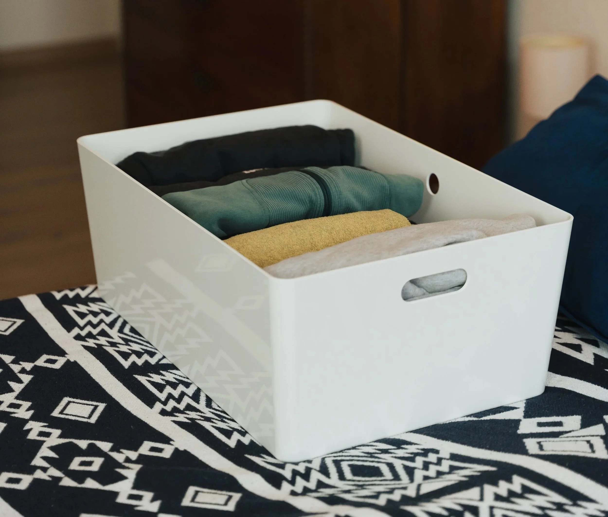 Weiße ordentliche Aufbewahrungsbox, gefüllt mit Kleidung in verschiedenen Farben, auf einer schwarzen Decke mit weißen Mustern.
