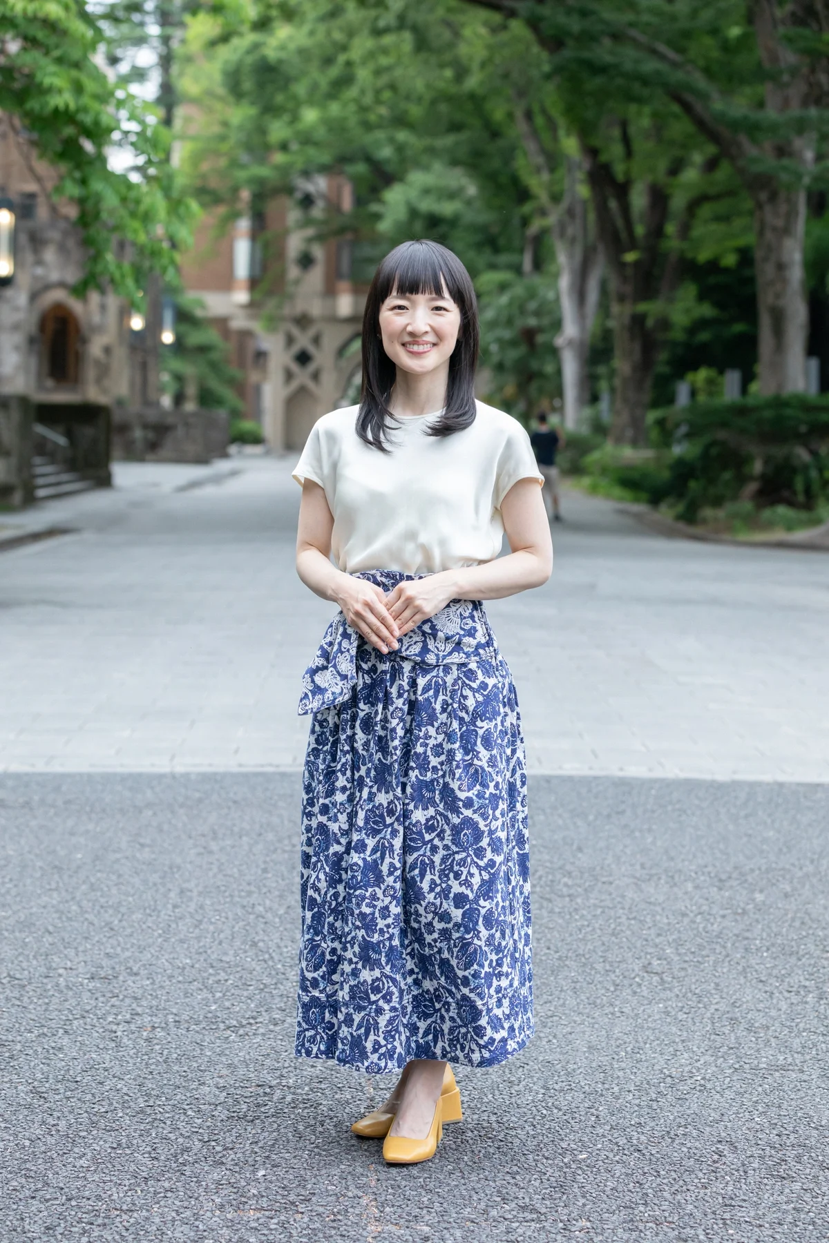 Marie Kondo, umgeben von grünen Bäumen und Gebäuden, trägt weiße Bluse, blaue gemusterte Hose und gelbe Schuhe.