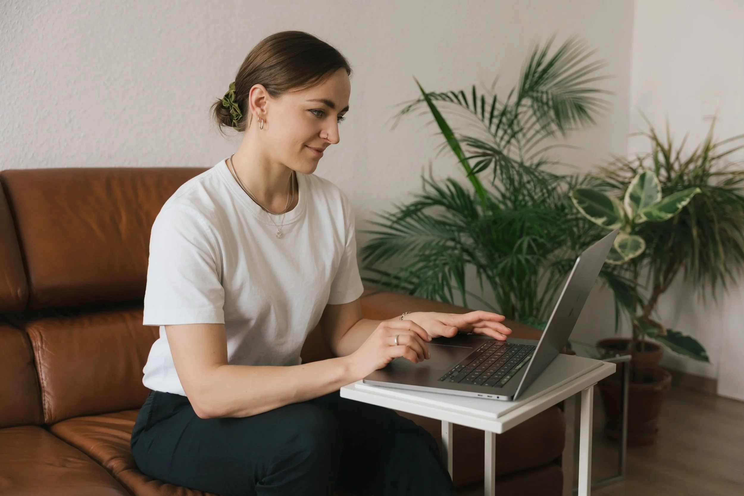 Antonia Grüner, Ordnungscoach, sitzt auf einer Couch, tippt auf einer Laptop-Tastatur, umgeben von grünen Zimmerpflanzen.