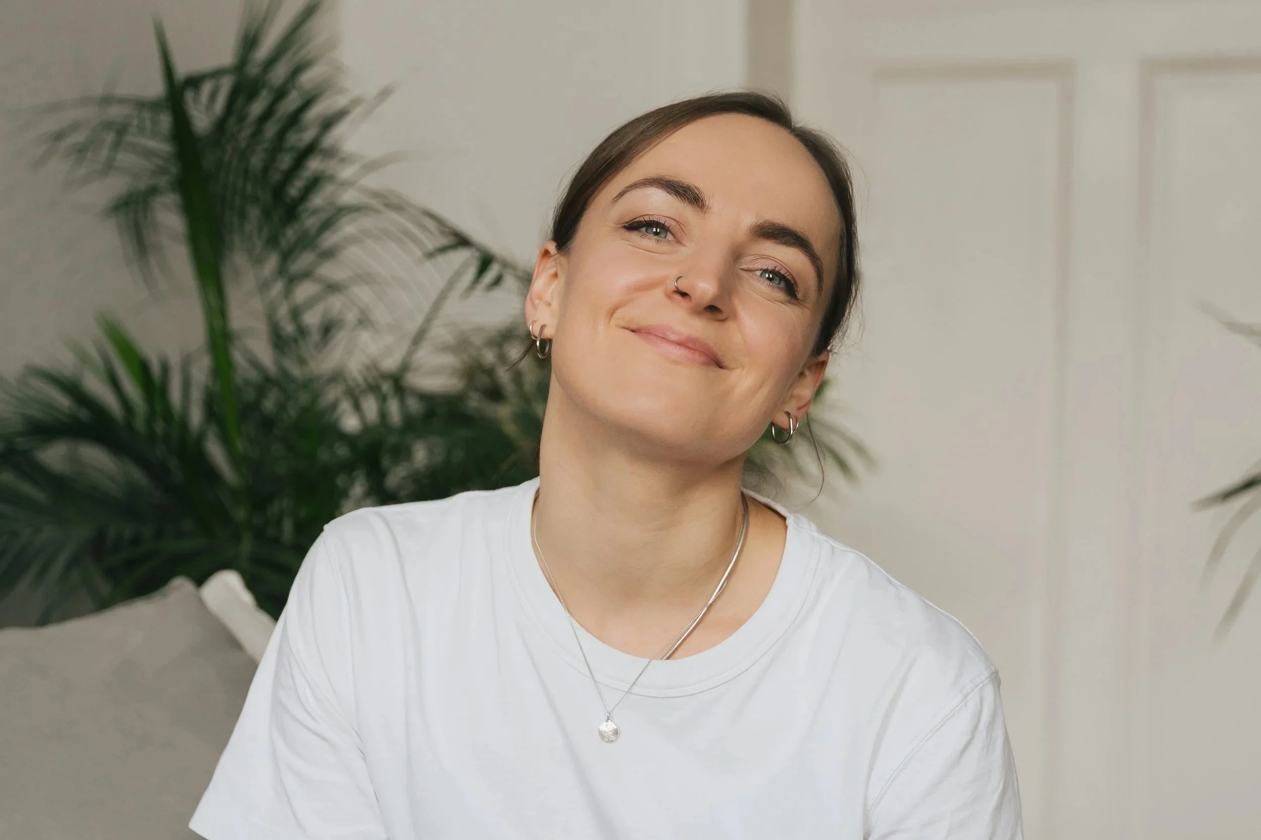 Antonia Grüner, Ordnungscoach, trägt ein weißes T-Shirt, lächelt mit geneigtem Kopf vor grüner Pflanze, weißem Hintergrund.