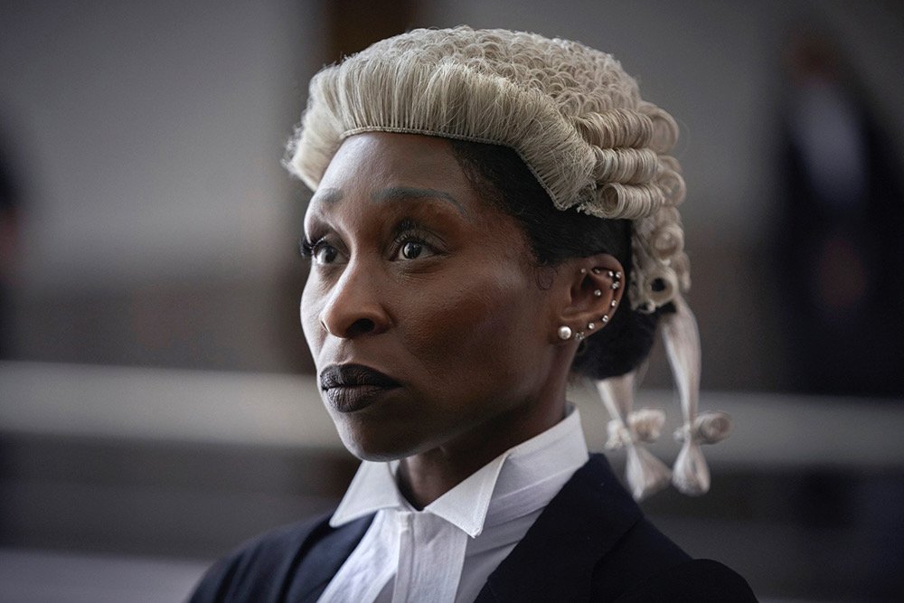 A woman with styled gray and black hair wearing a white court wig, black robe, and white collar, with multiple earrings.