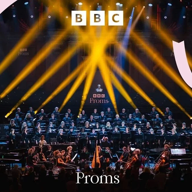 A large choir and orchestra on stage during a performance, with bright yellow and white stage lights and the BBC logo displayed overhead. The word 'Proms' is visible at the bottom of the image.