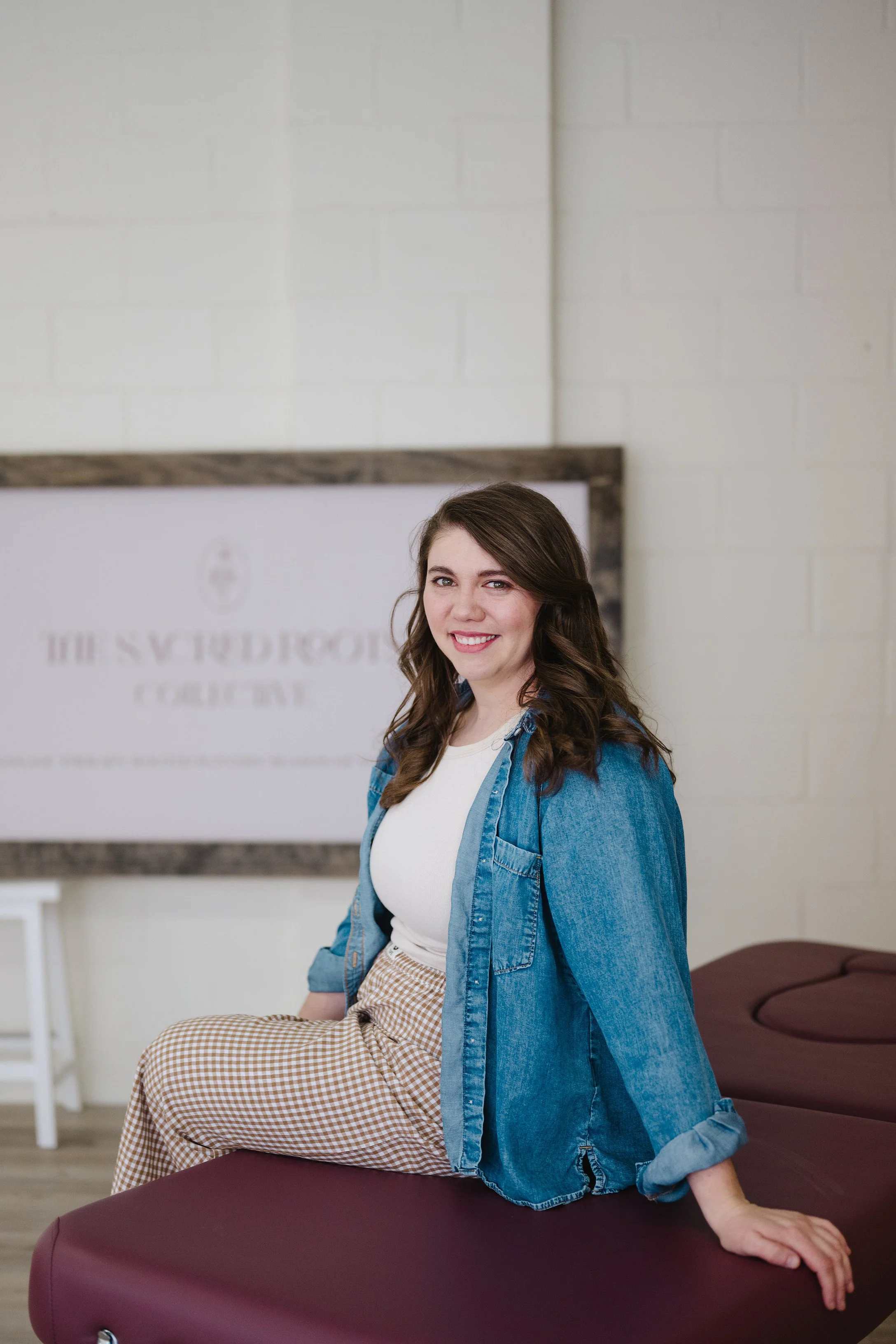 A young woman with long brown hair, wearing a white shirt and denim jacket, sitting on a massage table, smiling at the camera, in a wellness or therapy center.