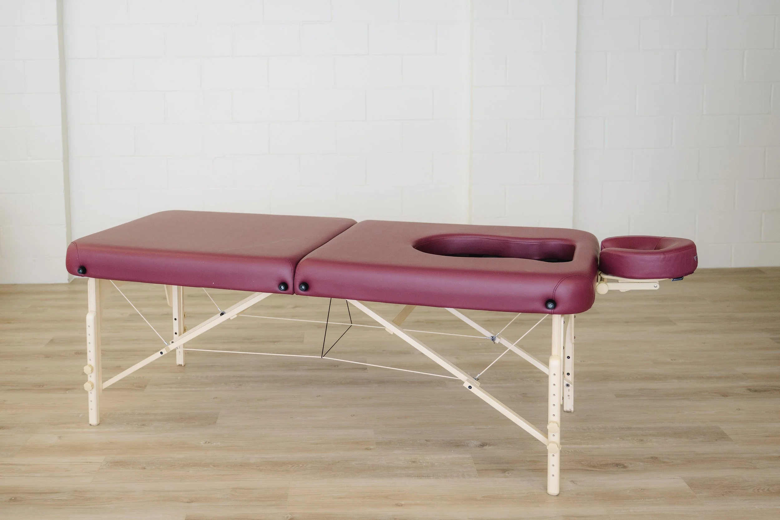 A massage table with burgundy padding on a wooden floor against a white brick wall.