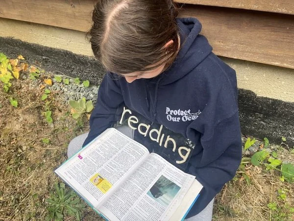 A girl in a navy blue hoodie sitting outside near a wooden wall, reading a textbook with the word "reading" spelled out in white clay on her hoodie.