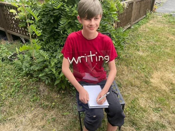 A boy with brown hair and a red shirt sitting outdoors on a small chair, writing in a notebook, with green bushes and a wooden fence behind him. "Writing" is spelled in white clay on his shirt.