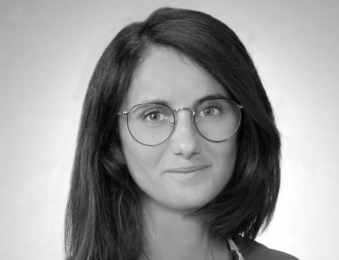Black and white portrait of a woman with straight, shoulder-length dark hair, wearing round glasses, and looking at the camera with a slight smile.