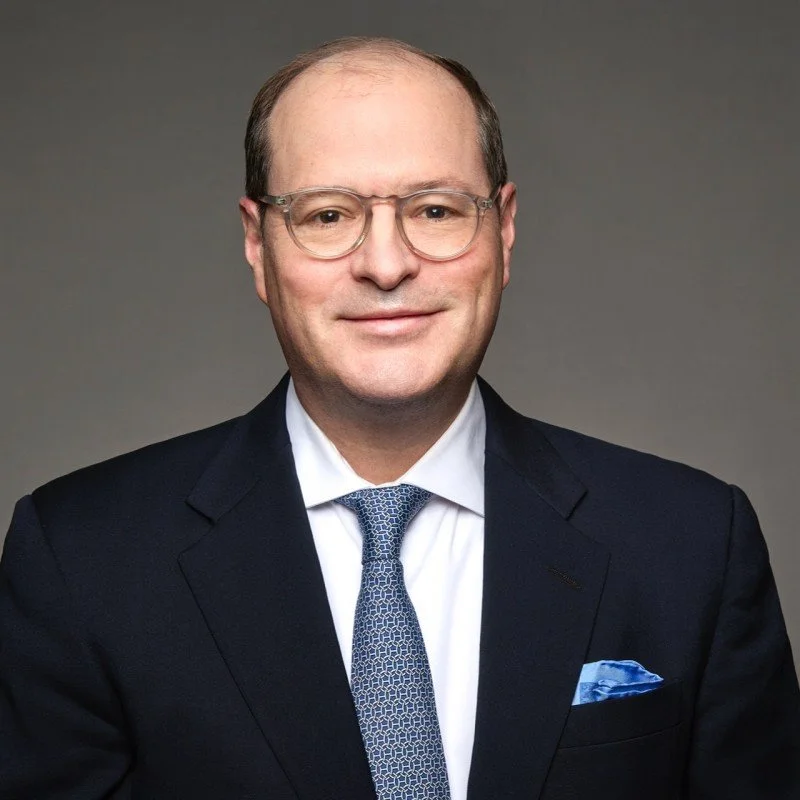 A middle-aged man with short light brown hair, glasses, wearing a dark suit, white shirt, and patterned blue tie, smiling against a gray background.