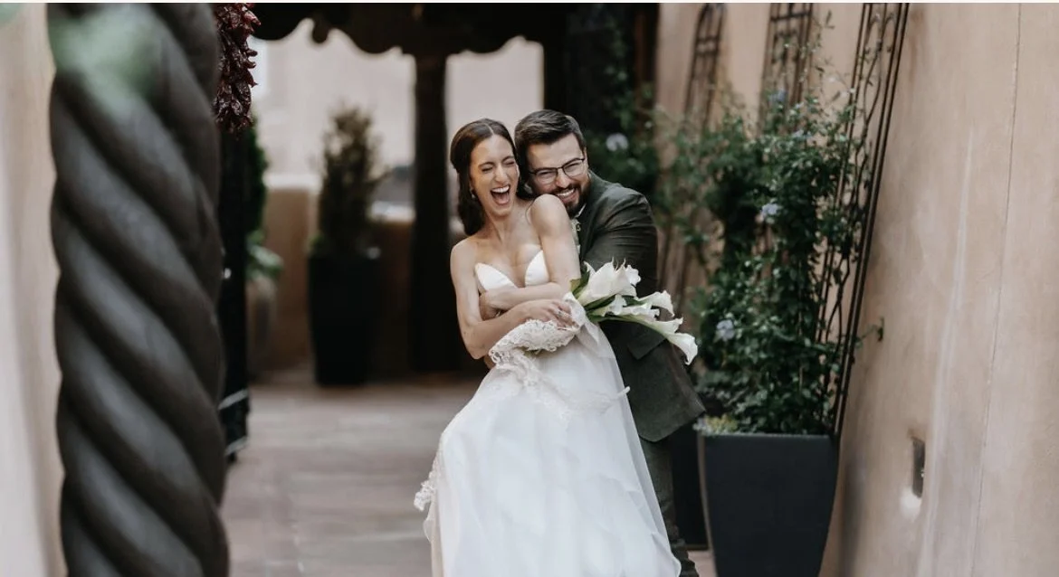 A joyful bride and groom sharing a hug and laugh in a romantic outdoor setting, with greenery and a stone wall in the background.