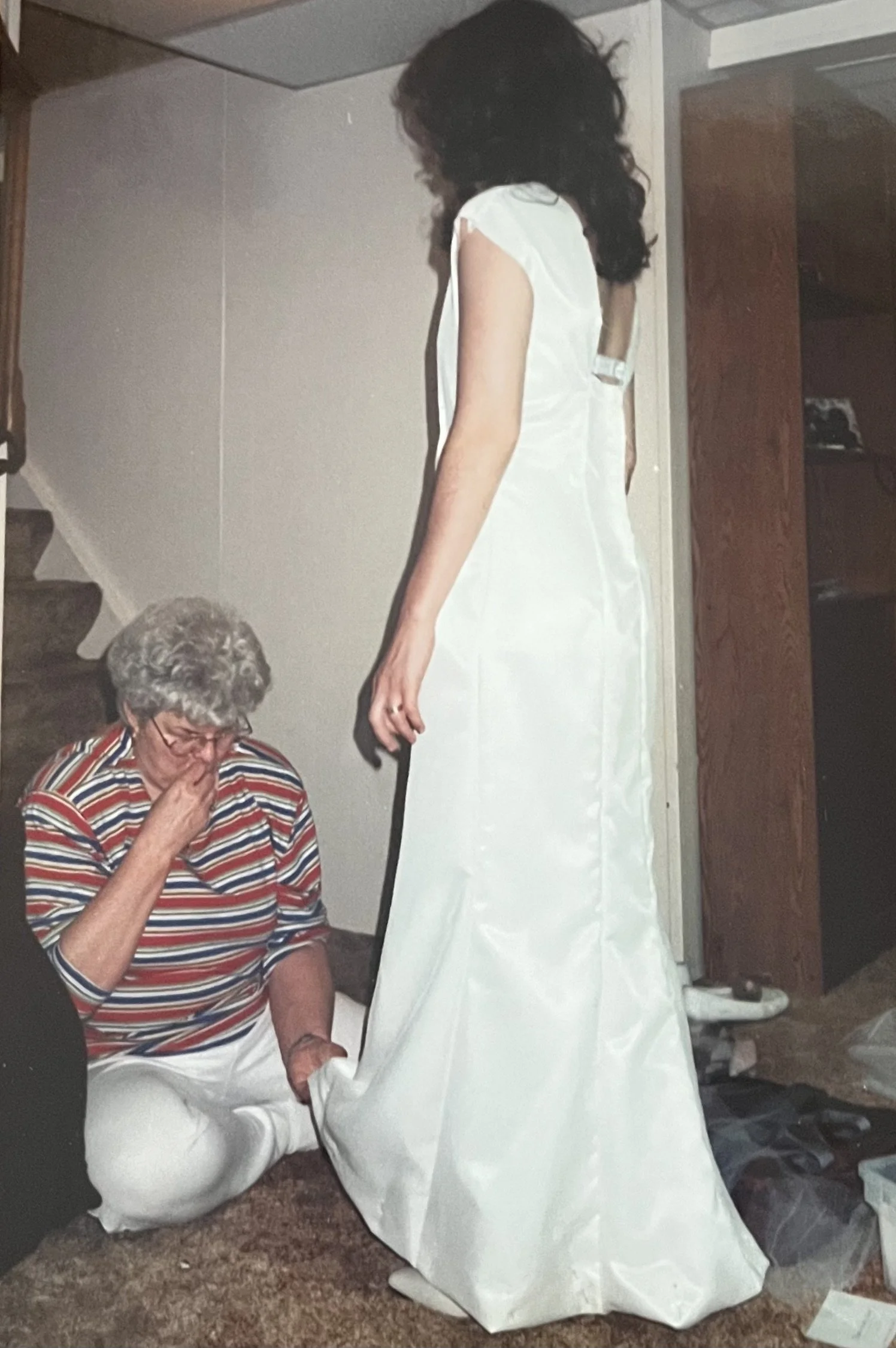 A woman with gray, curly hair and glasses is sitting on the carpeted floor, appearing emotional, with her hand covering her nose and mouth. She is wearing a striped shirt and white pants. Standing beside her, a young woman with long, dark, curly hair, dressed in a white satin wedding gown, is looking down at her.
