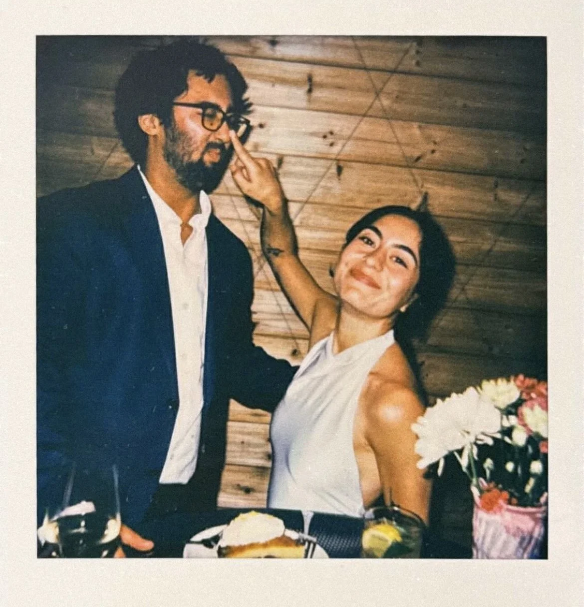 A man and woman are smiling at a celebration. The man, wearing a dark suit and glasses, has a beard and is touching his glasses. The woman, in a white sleeveless dress, has her arm raised playfully toward the man's face. There are flowers on the table and a wooden wall in the background.