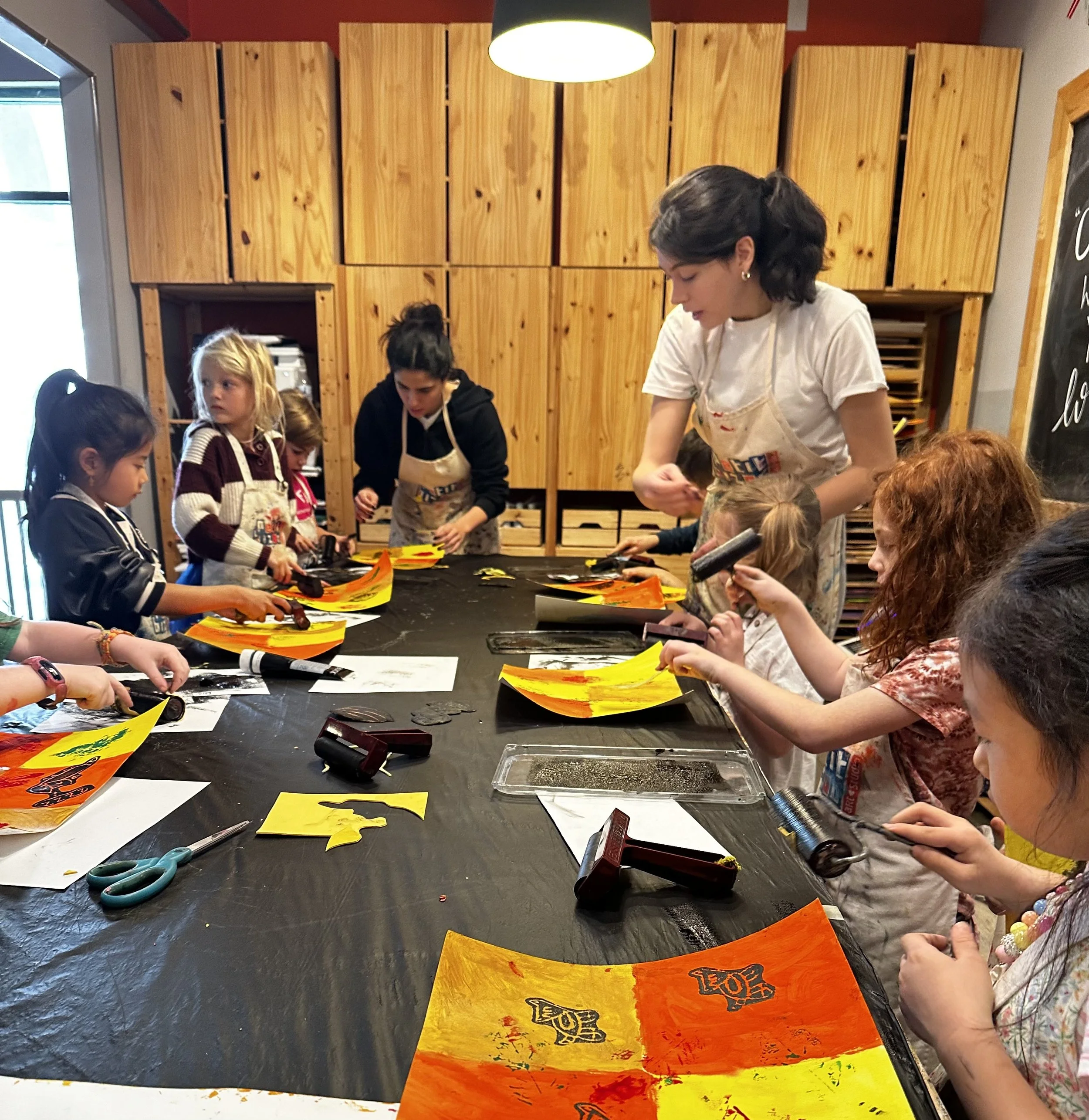 Children and an adult at a long table in a wood-paneled room, preparing clay for a pottery activity, with tools and supplies scattered on the table.