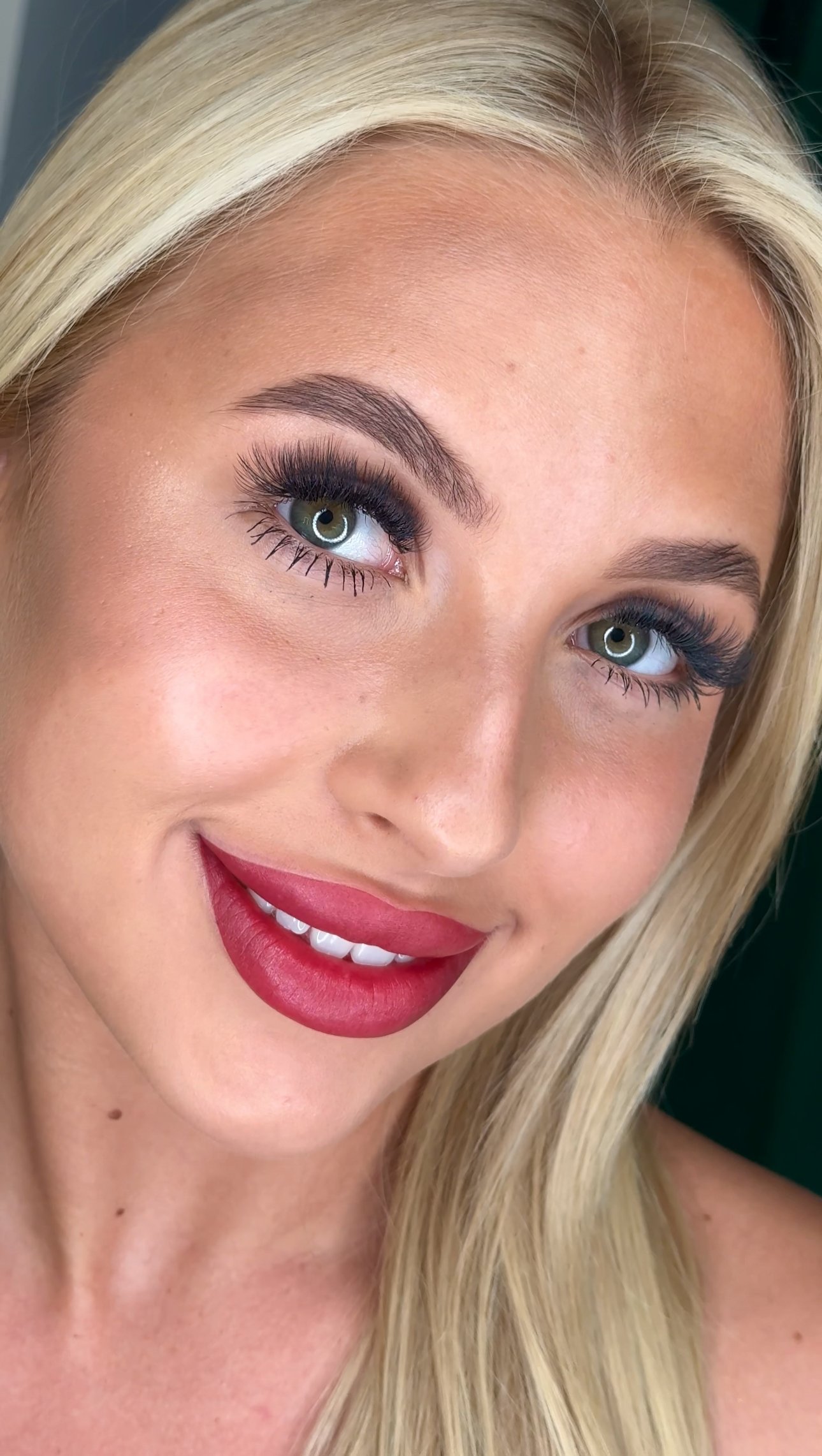A smiling woman with long blonde hair, green eyes, makeup including dark eyelashes, and red lipstick.
