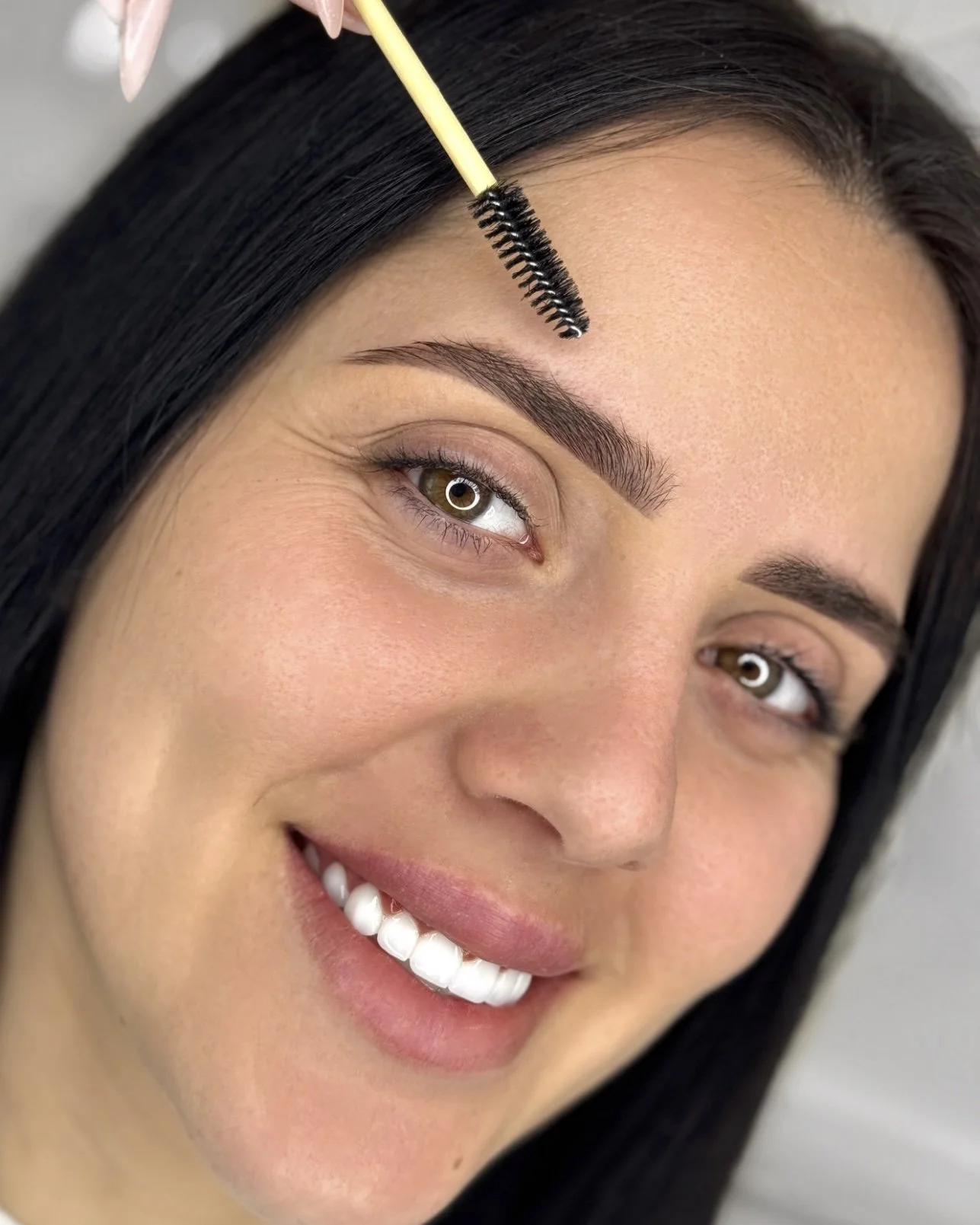 Close-up of a woman with dark hair smiling, as a person applies eyelash extensions with a spoolie brush.