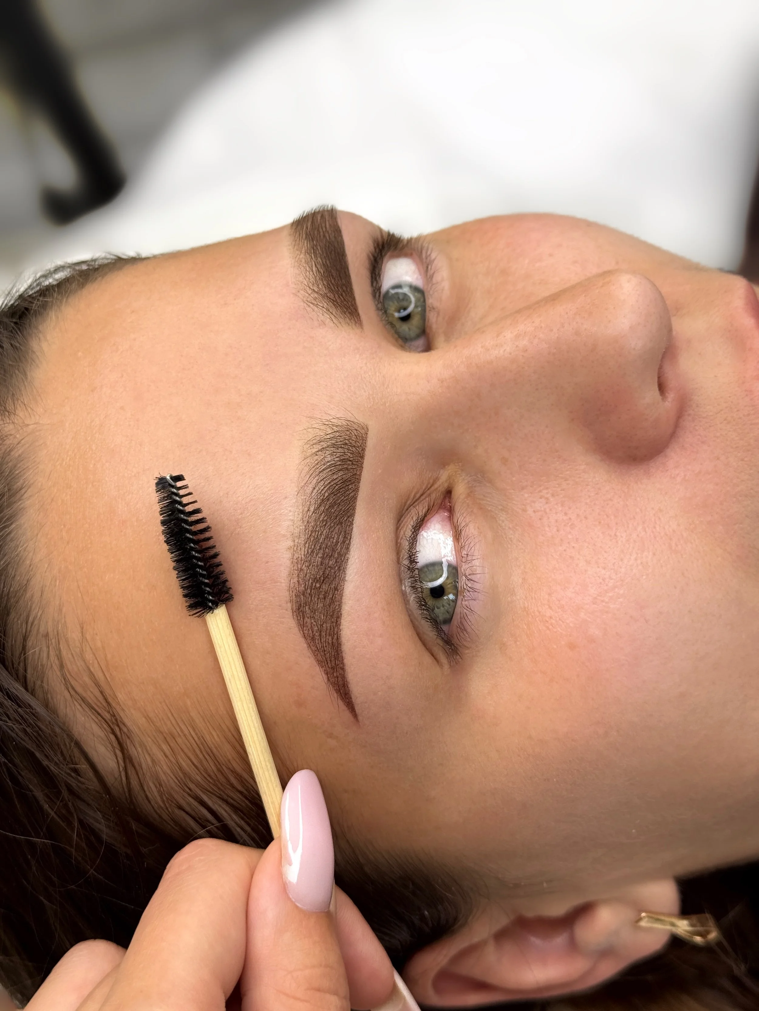 Close-up of a woman with freshly made-up eyebrows, looking upwards, while a makeup artist applies mascara to her eyebrows with a spoolie brush.